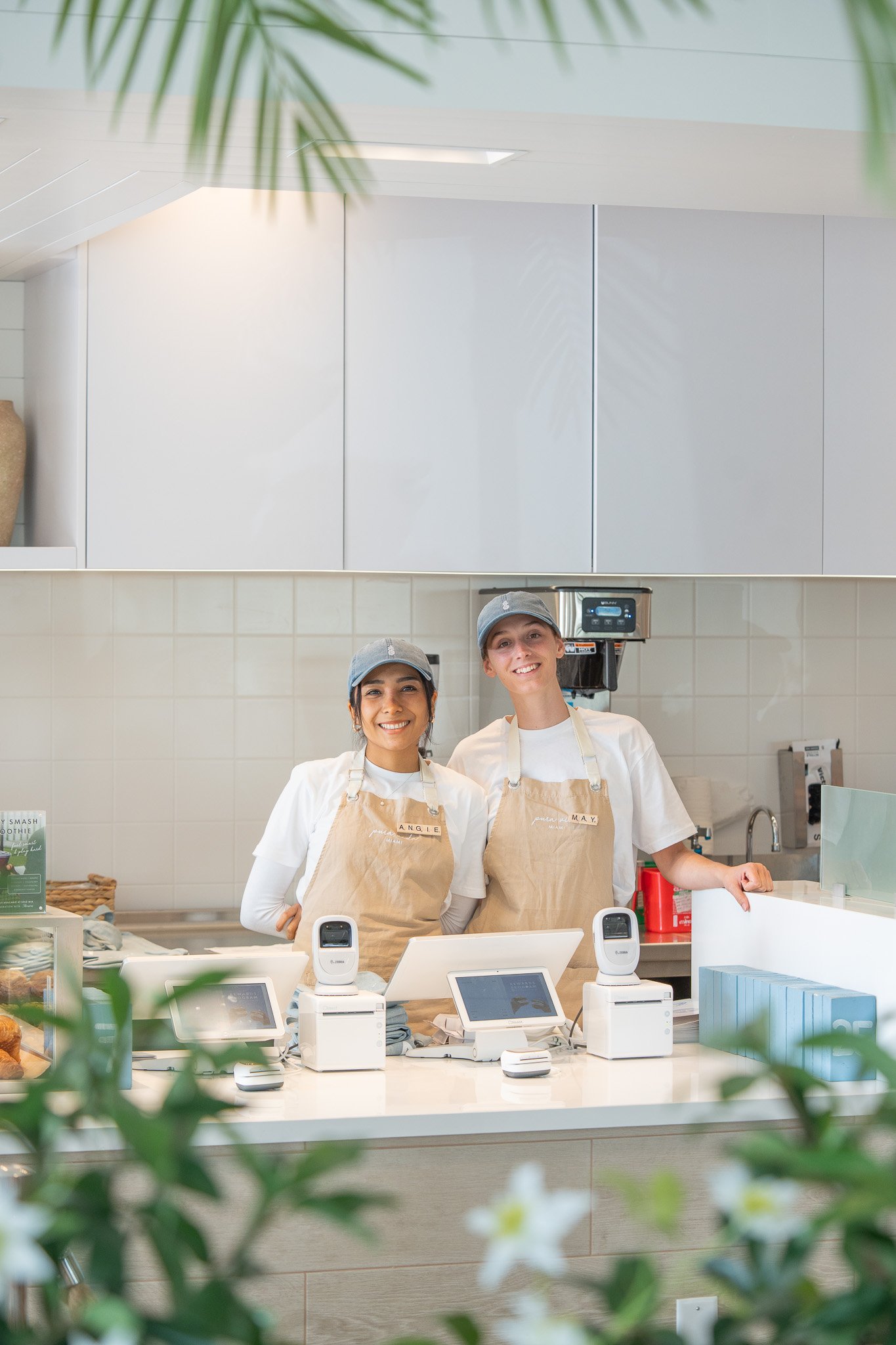 Two smiling staff members wearing aprons and hats, standing behind a white counter with digital registers, in a bright, modern cafe setting with plants in the foreground.
