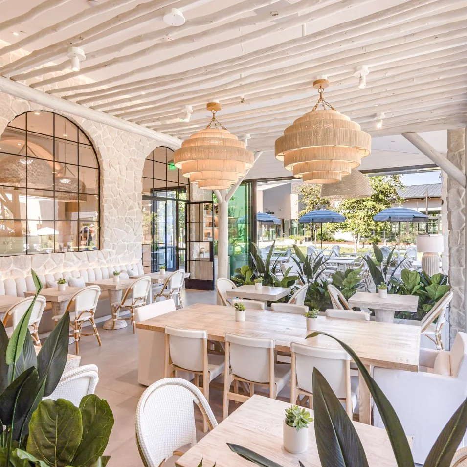 Bright and airy restaurant interior with light wood tables, white chairs, large green plants, stone walls, and hanging beige pendant lights. Large windows and outdoor seating with umbrellas visible.