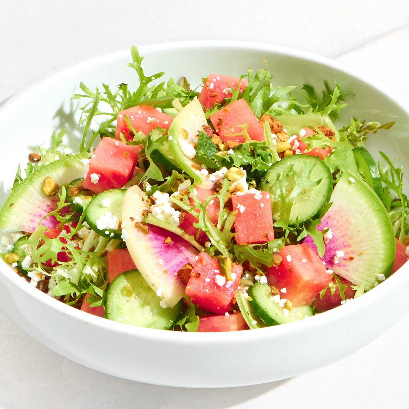 Fresh salad with cucumbers, watermelon, radish slices, arugula, and feta cheese in a white bowl.