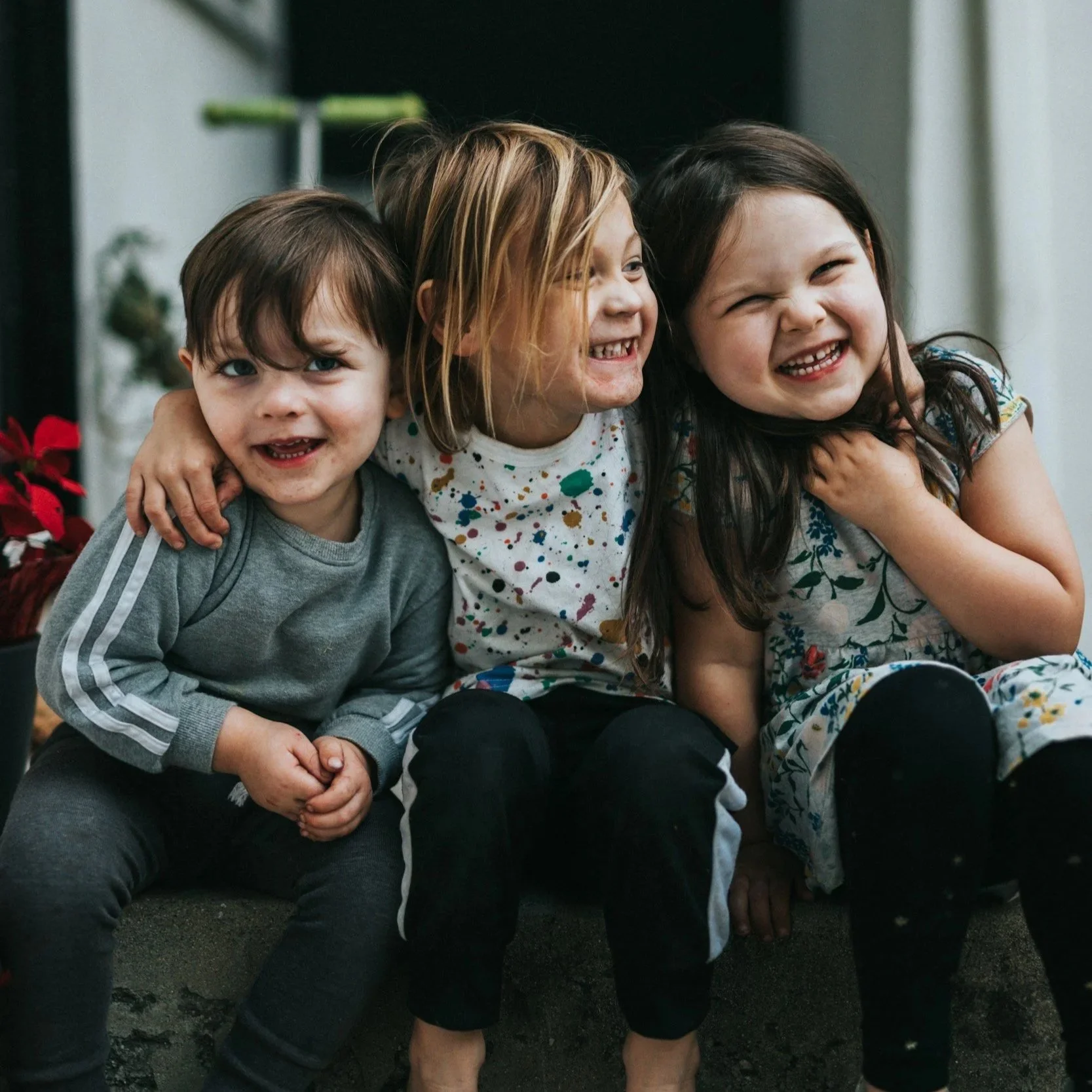 Three children sitting closely together, smiling and laughing, outdoors.