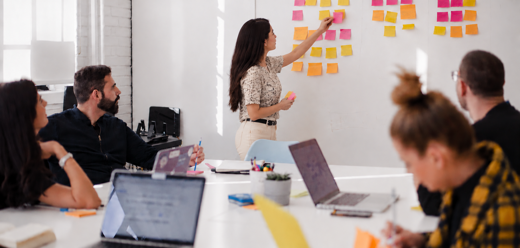 A woman presenting sticky notes on a whiteboard to a group of people in a meeting room.