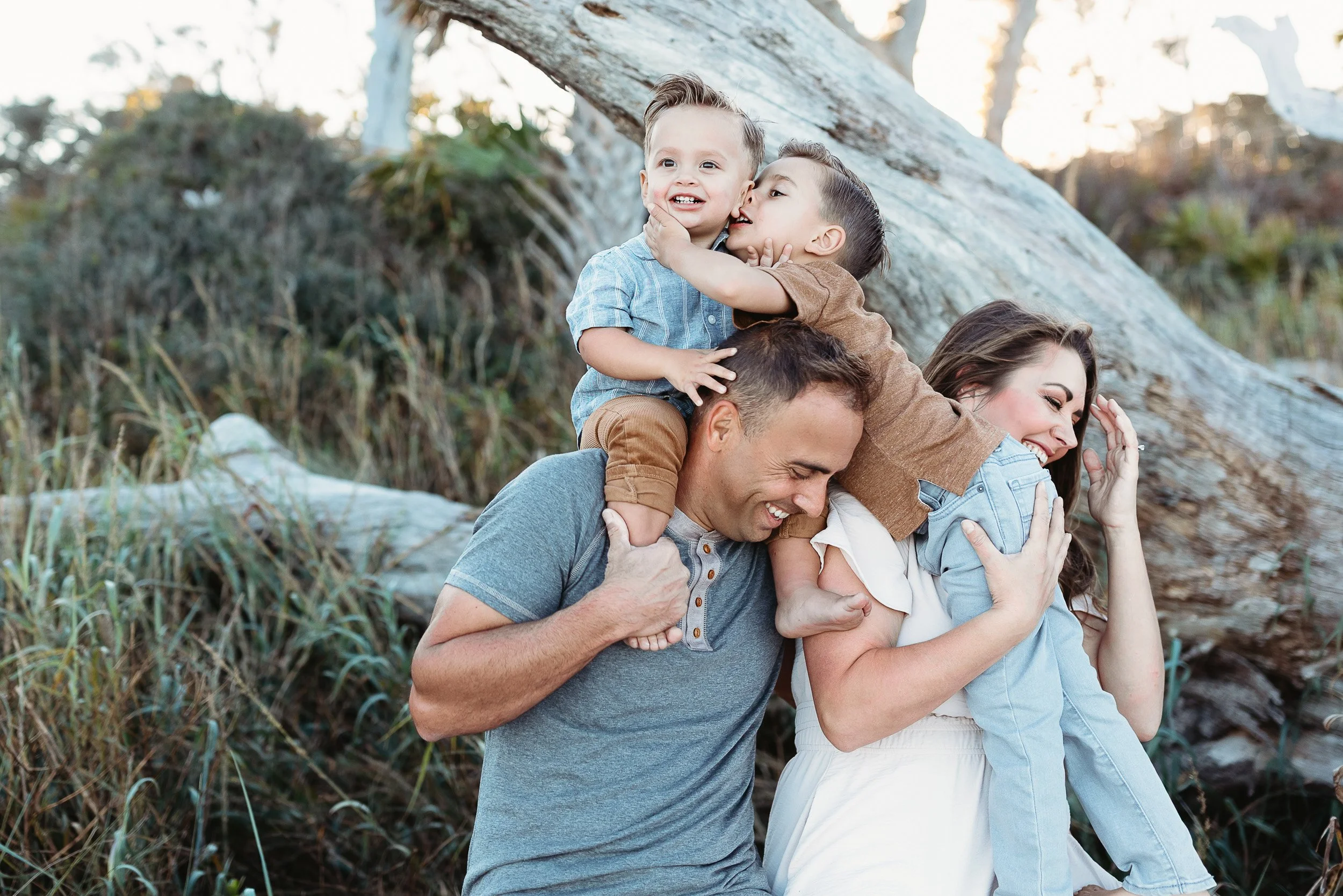 toddler boys on parent;s shoulders at the beach for family photos in Jacksonville beach boys are kissing and parents are laughing