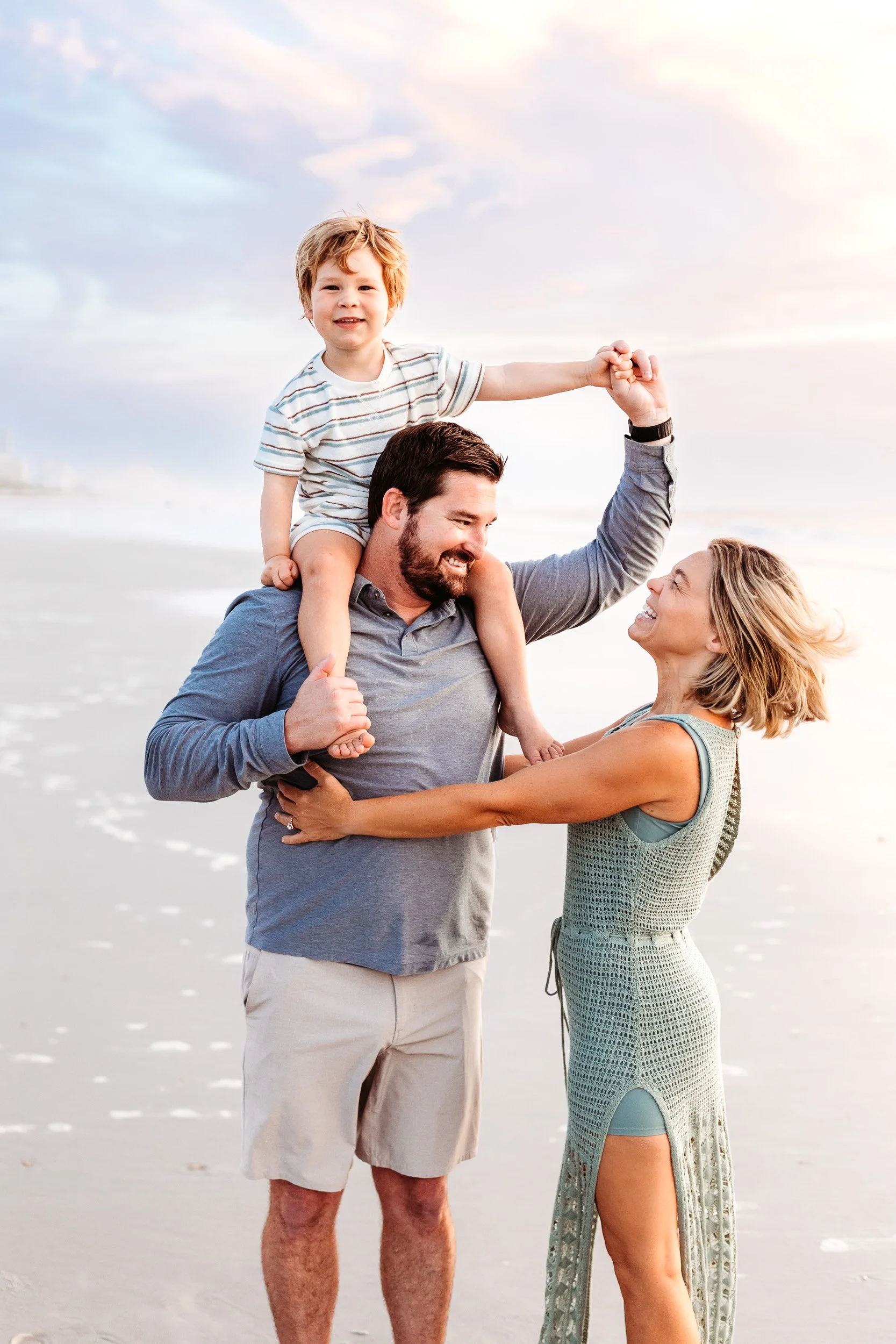 little boy on dad's shoulder and the family is laughing pastel pink sunrise sky behind them