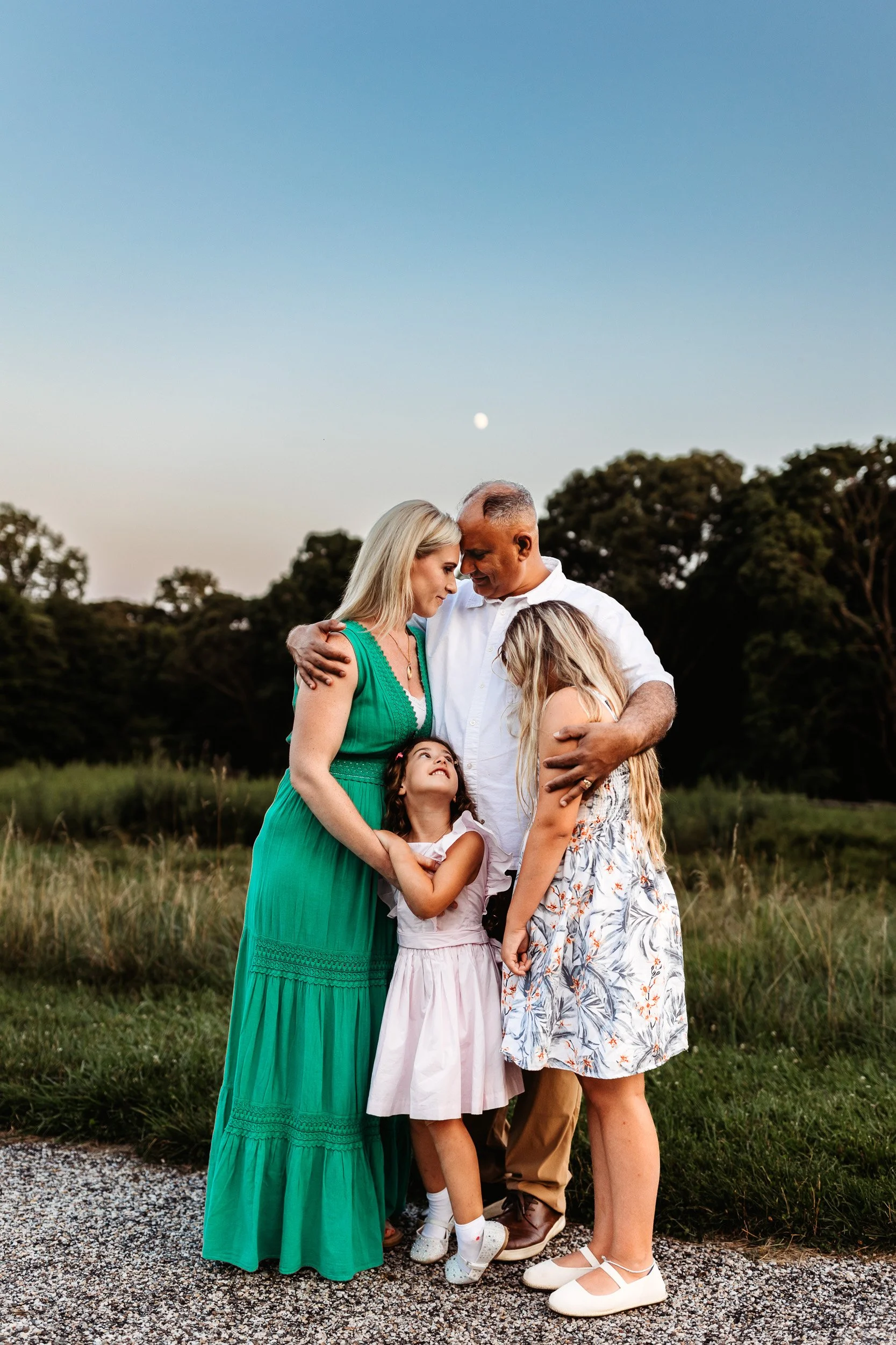 intimate image of a husband and wife embraced while they hold their daughters and the moon is shining above