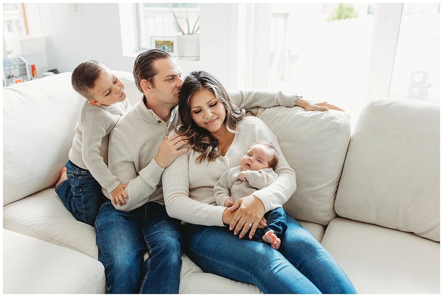 family seated on couch in jeans and tan sweaters dad kissing mom boy watching baby brother who mom is holding