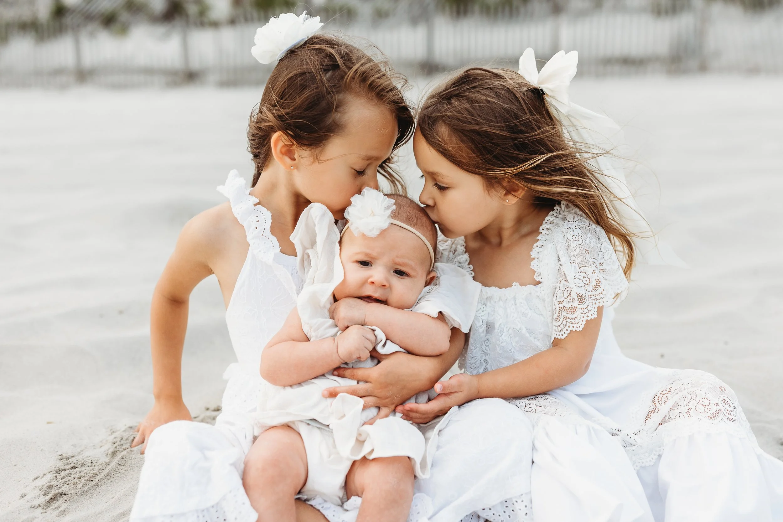 toddler sisters holding newborn baby and kissing her while seated on the beach at sunset