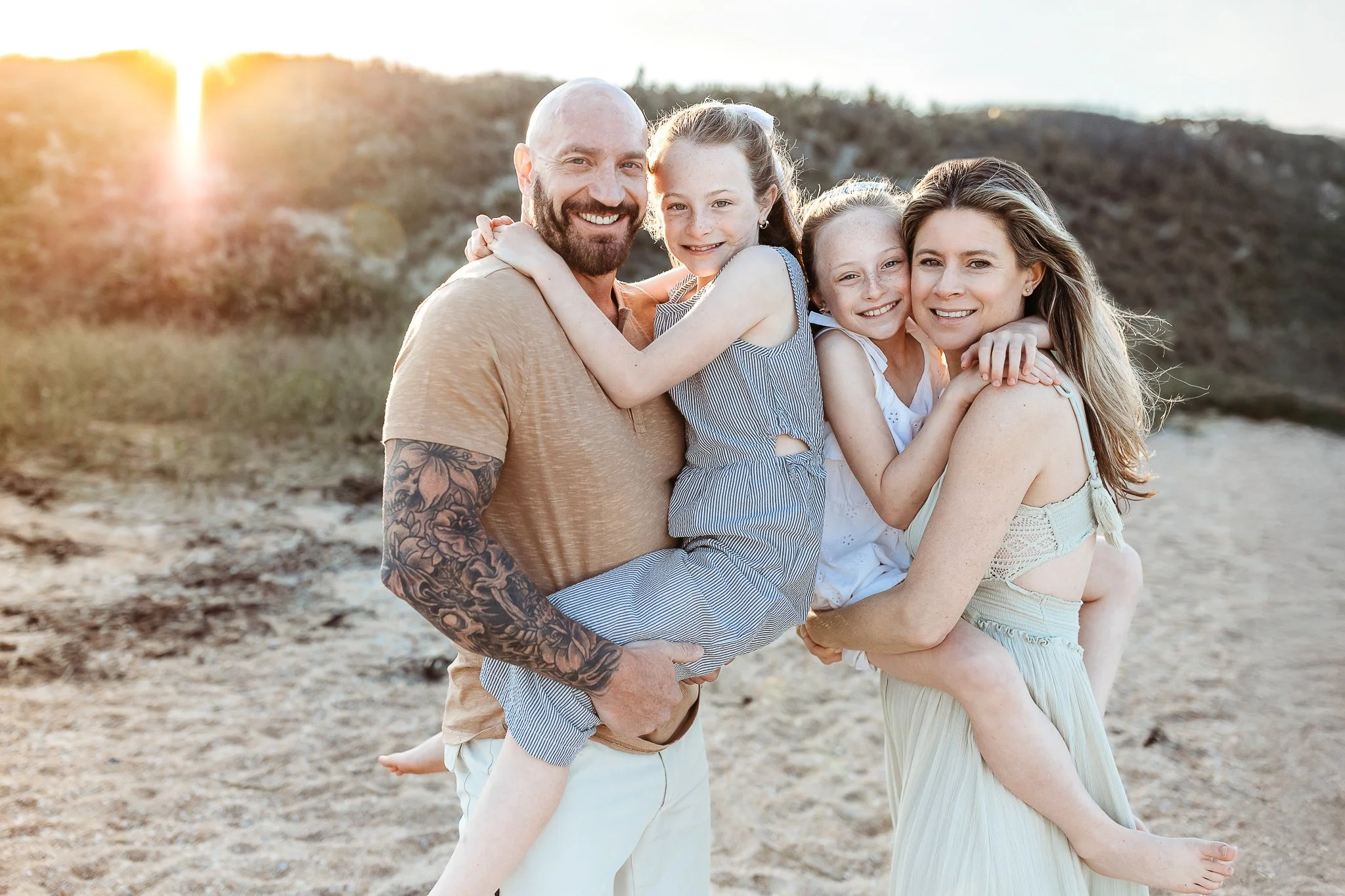 close up of a family on Neptune beach at sunset and they are holding their children and in front of a dune at sunset mom is in a free people dress 