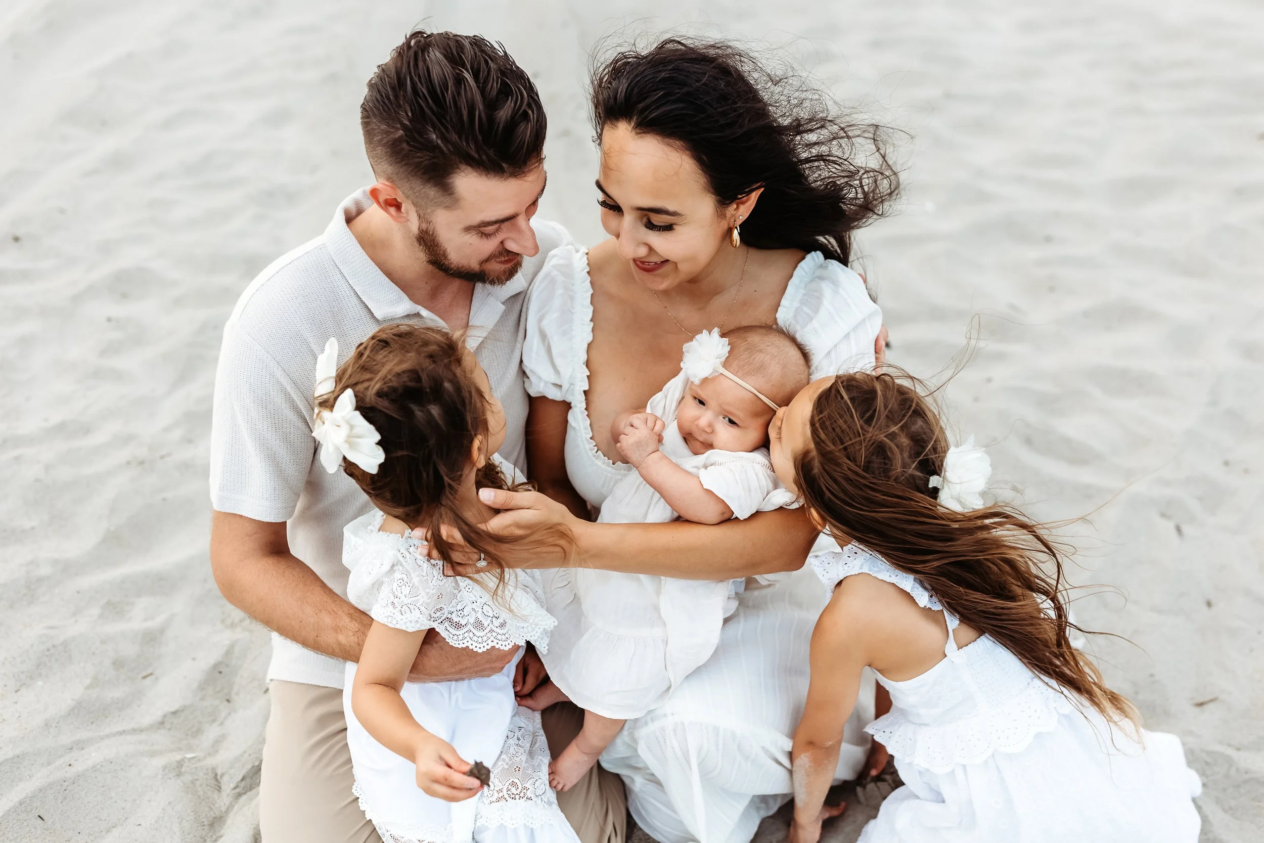 family seated on the sand and they are all looking at the new baby in mom's arms and they are dressed in white and tan