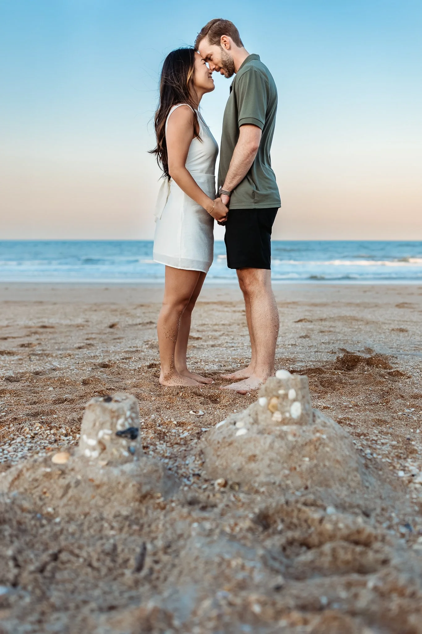 couple holding hands and leaning head to head in front of a sand castle at guana beach at blue hour with a pink and blue sky