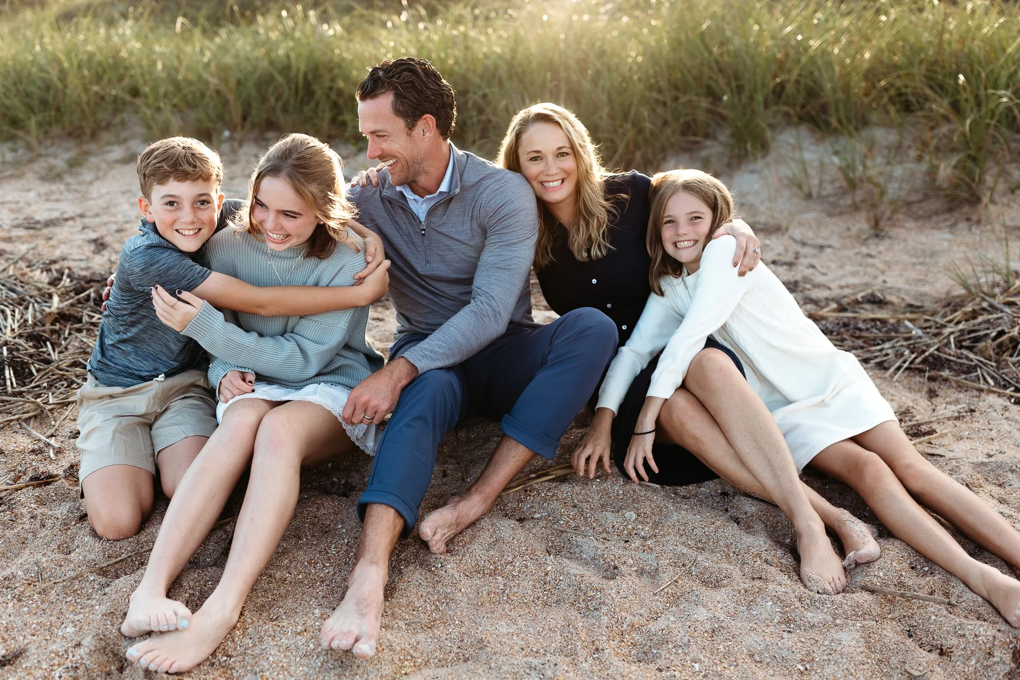 family embraced on the beach at sunset and they are all laughing while seated on the sand in front of a dune