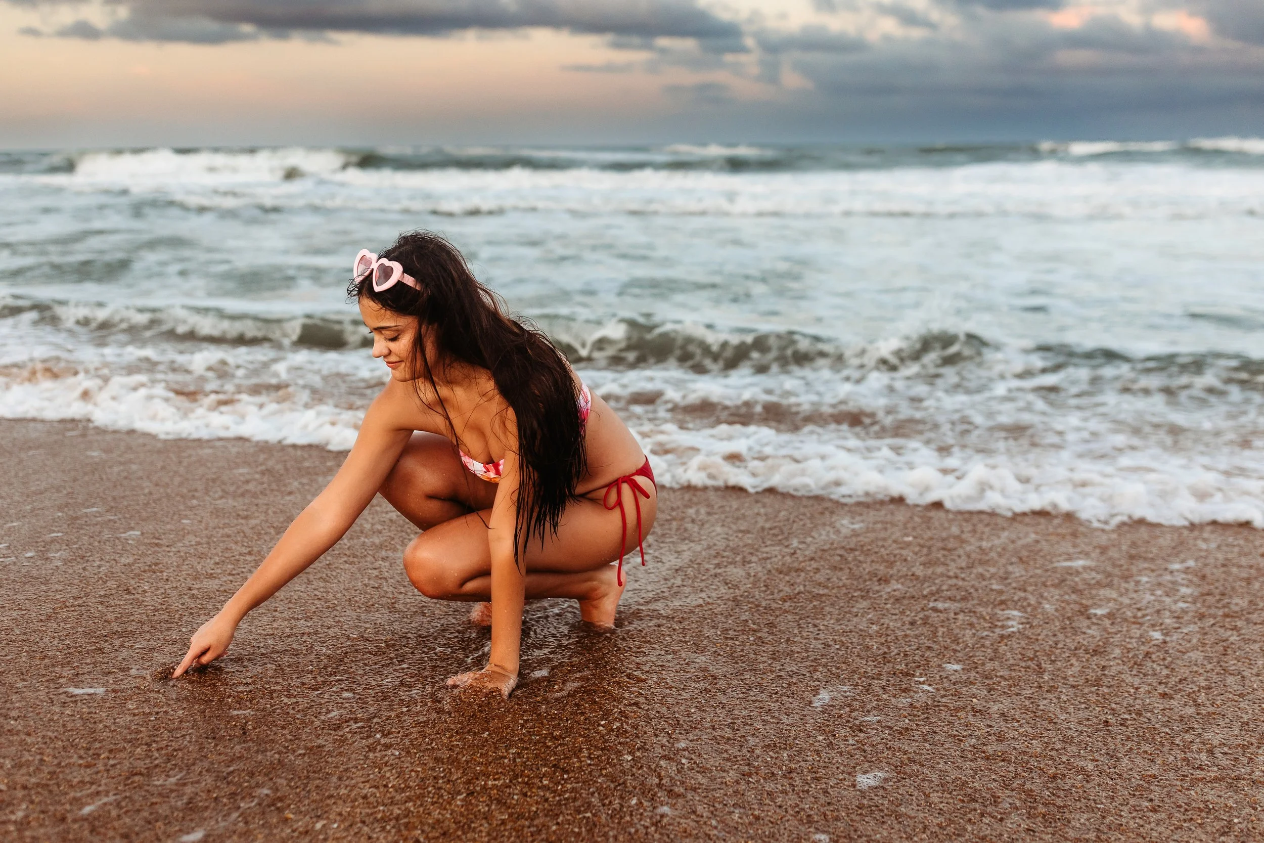 girl in bathing suit with heart glasses drawing in the sand at sunset
