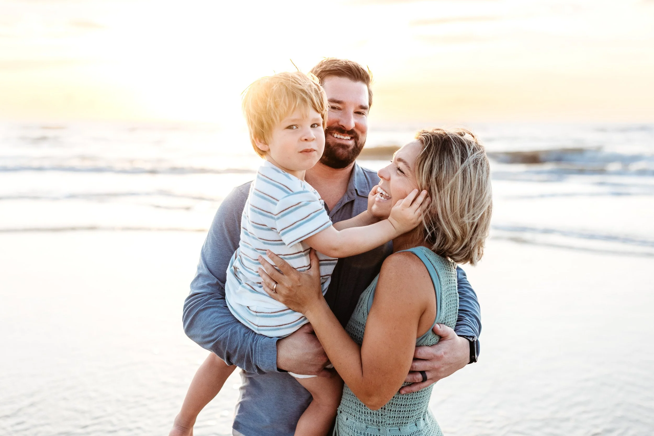 toddler boy grabbing mom's face and she is laughing and the sun is rising over the ocean behind them