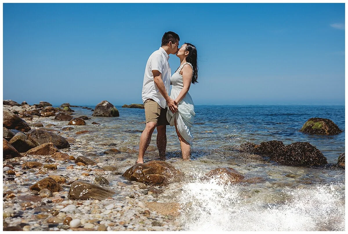 couple kissing on rocky beach while waves crash on boulders with bright blue sky behind them