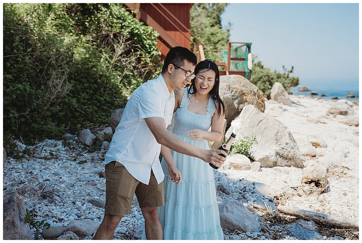 couple popping a bottle of veuve champagne on the rocky each and laughing while it fizzed over
