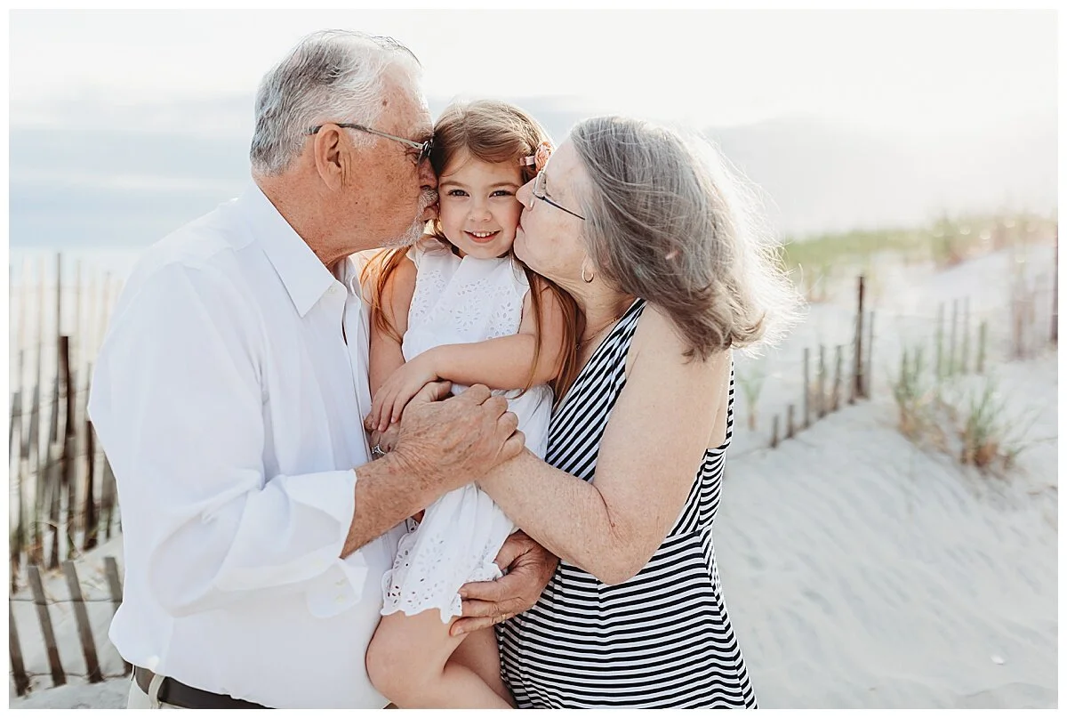 grandparents kissing granddaughter on the cheek on the beach in front of a dune while granddaughter laughs