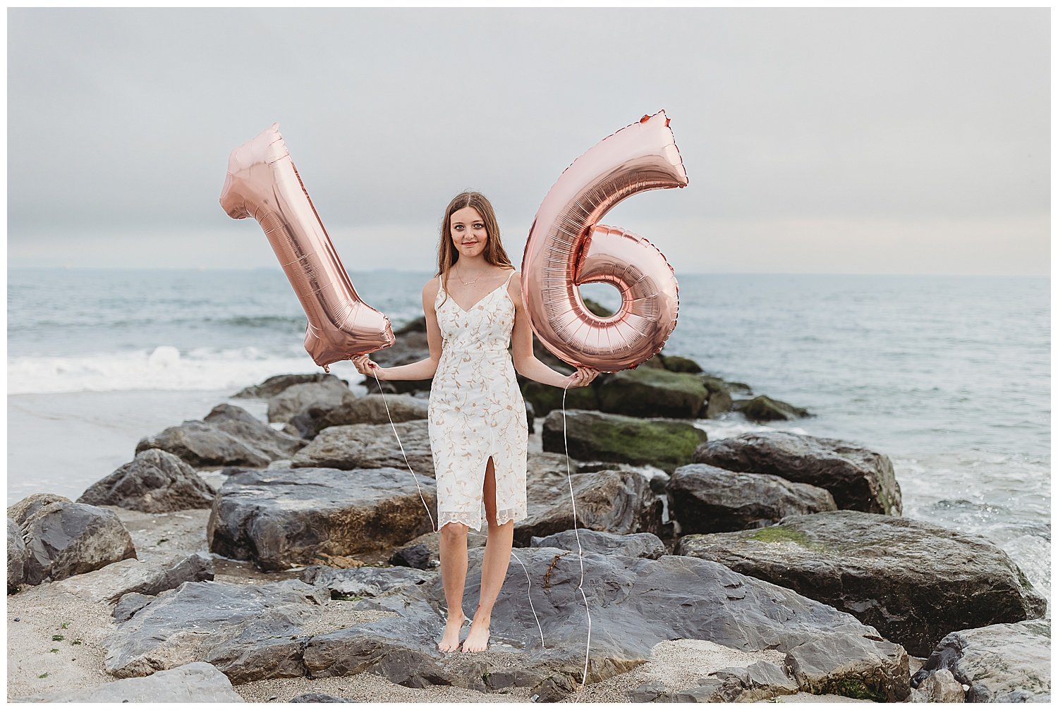 girl in white embroidered dress on a jetty with the ocean behind her holding 16 metallic balloons