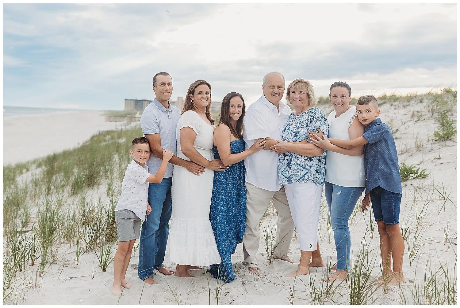 family dressed in white and blue with grandparents on a dune at the beach on a hazy cloudy evening embraced