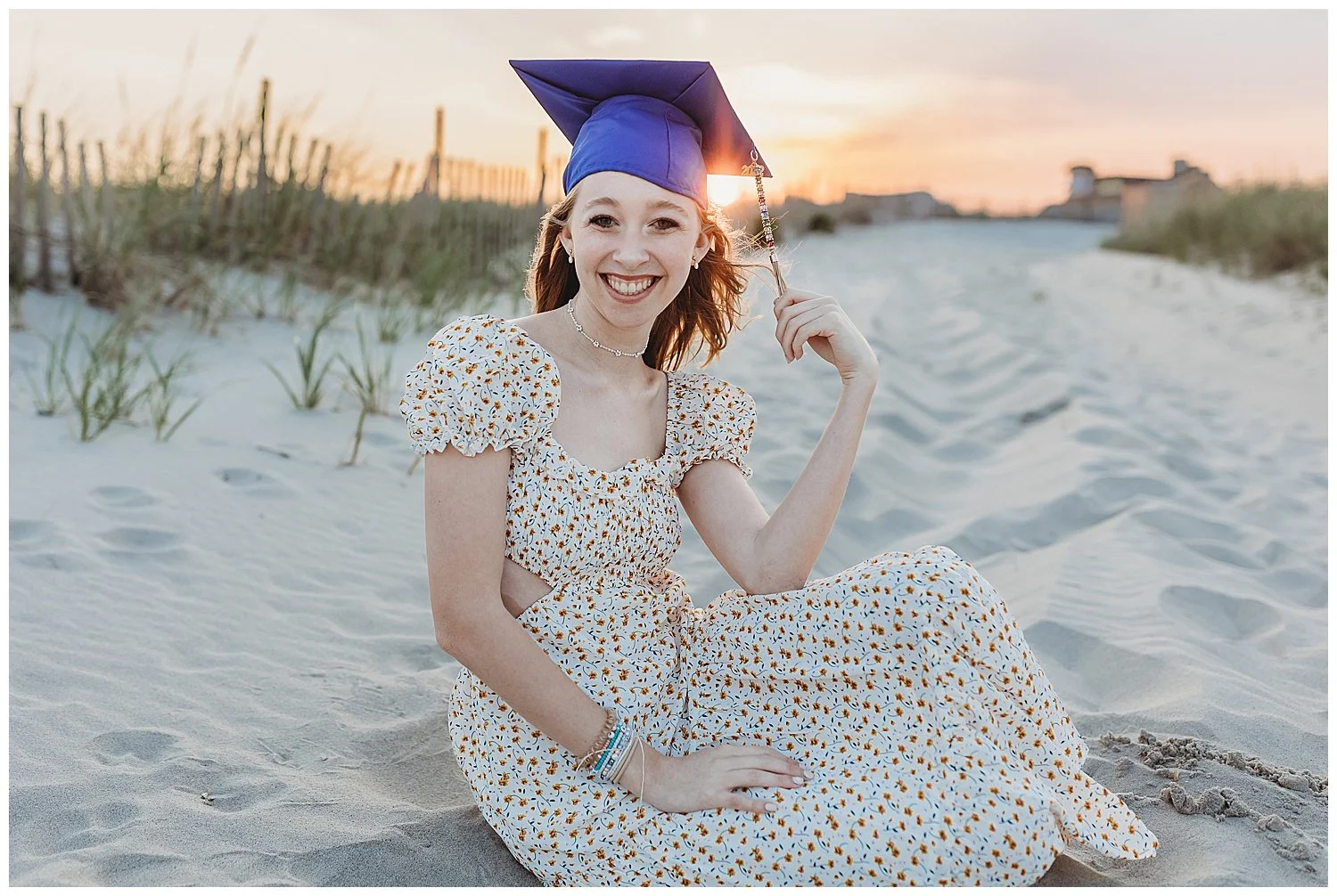 girl seated on Jacksonville beach at sunset with sunlight glowing through her cap and gown as she holds the tassel