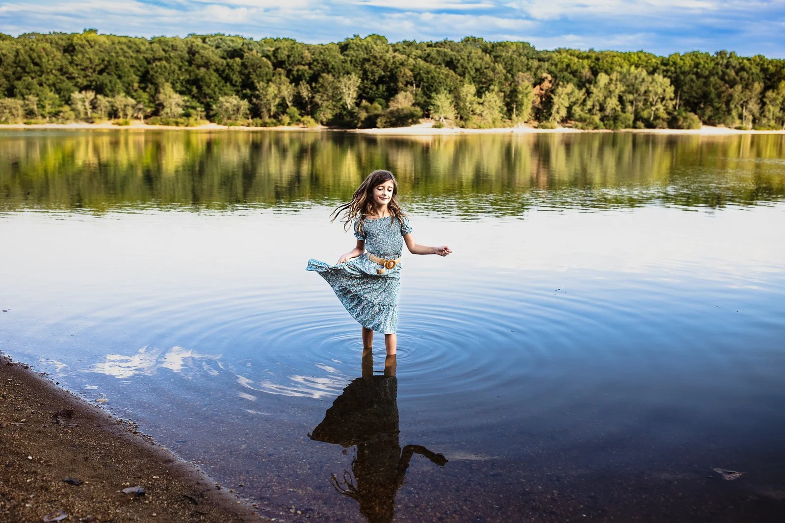 girl dancing in a lake ith greens and bleu sky behind her with a reflection in the water
