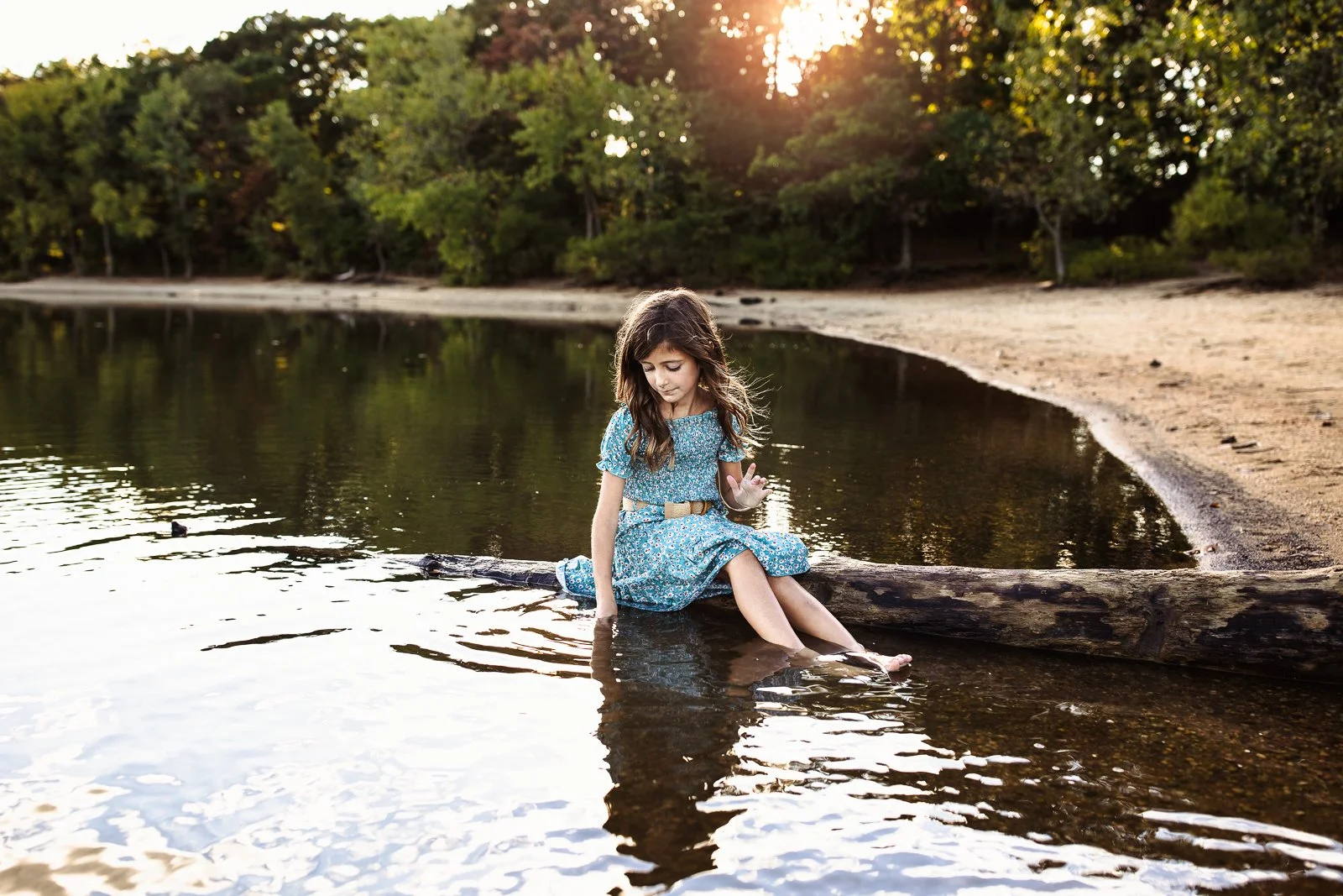 girl in bohemian maxi dress seated on a log at sunset in a lake playing with the water