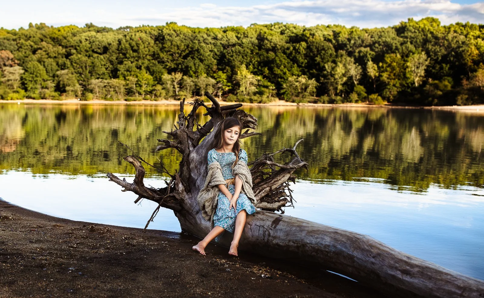 girl in maxi dress and sweater seated on driftwood at sunset in a lake
