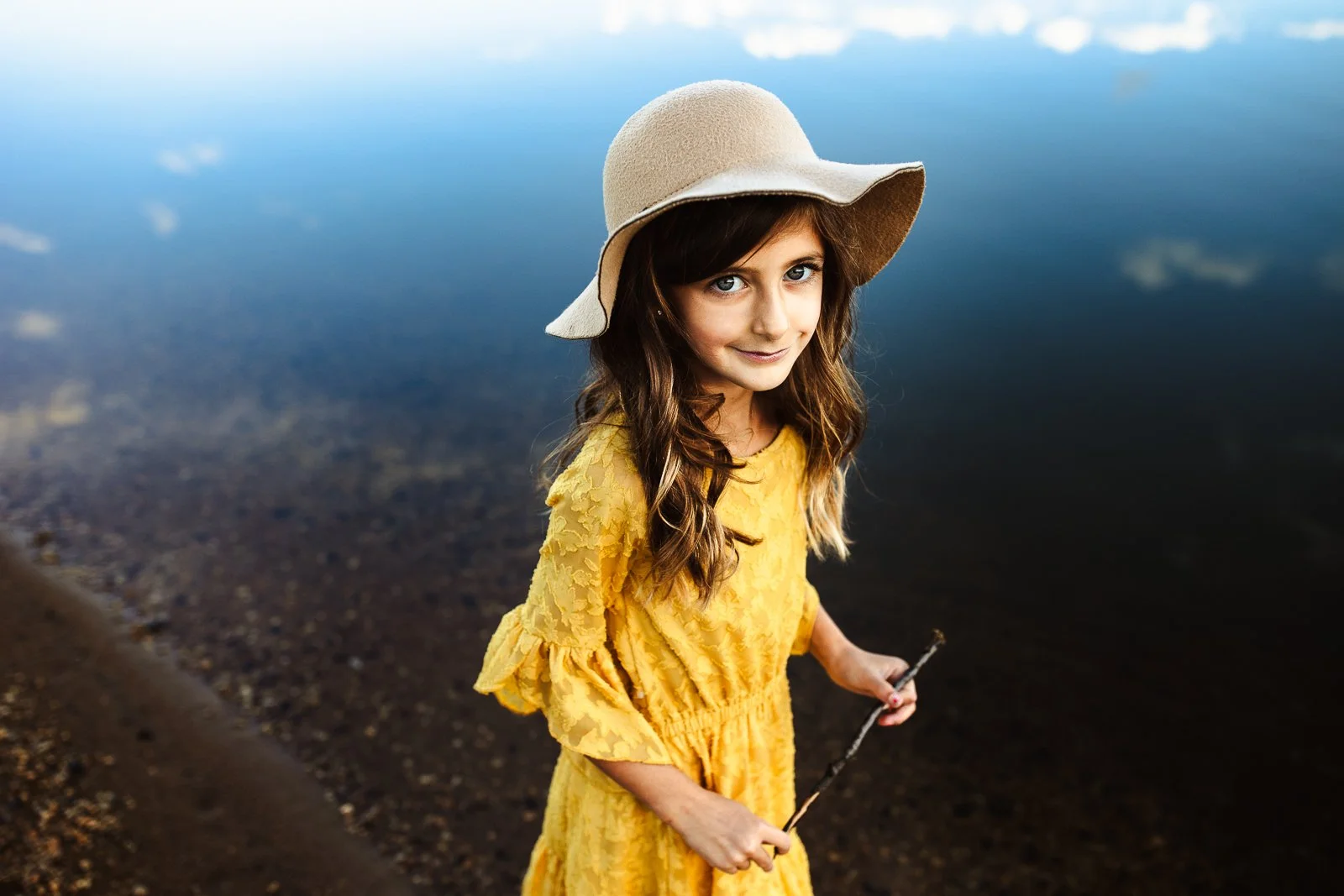 close up of young girl with floppy hat and yellow dress in a lake with reflection of clouds around her