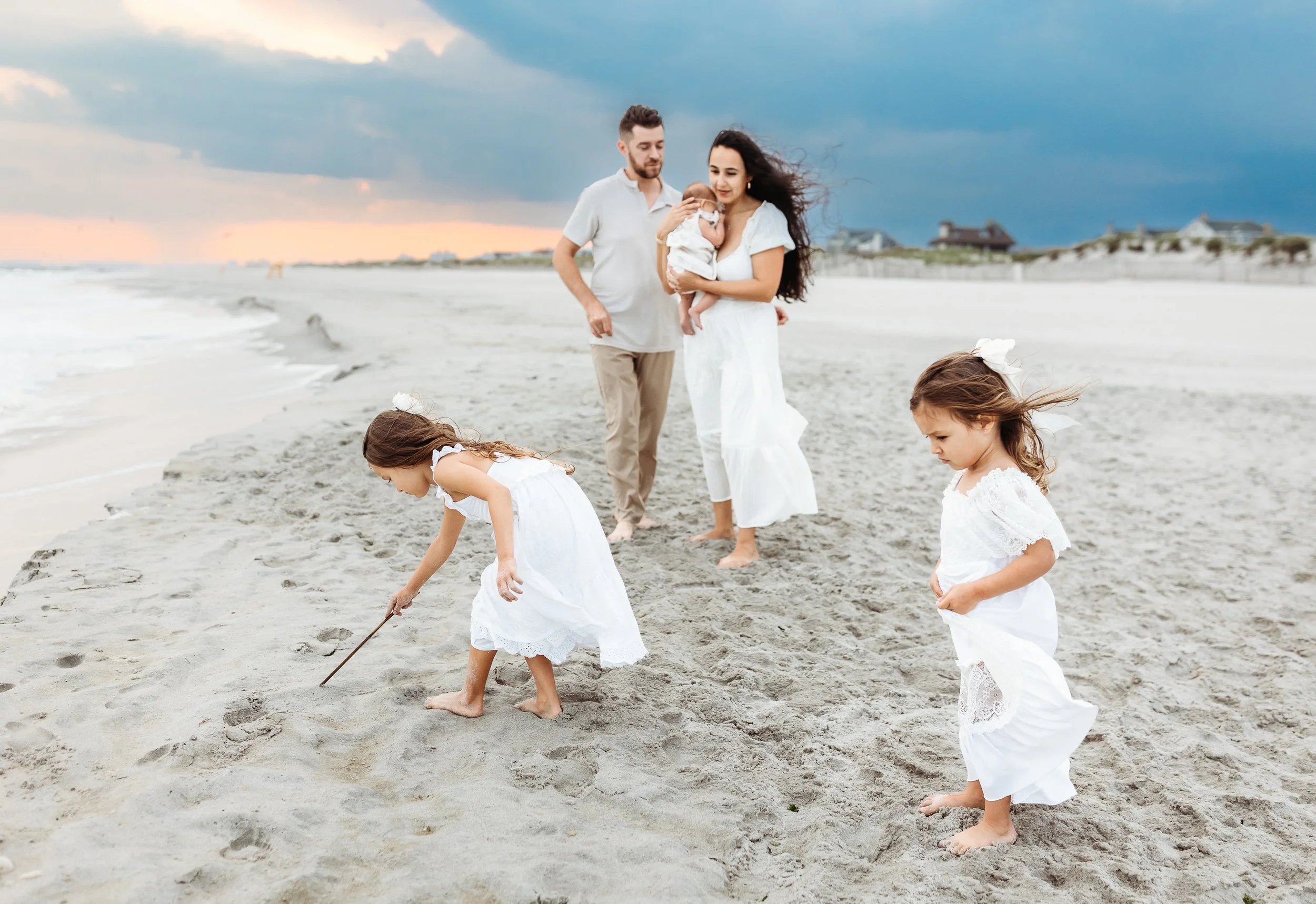 toddlers playing in the sand at the Atlantic beach and parts watching in the background while holding a newborn baby