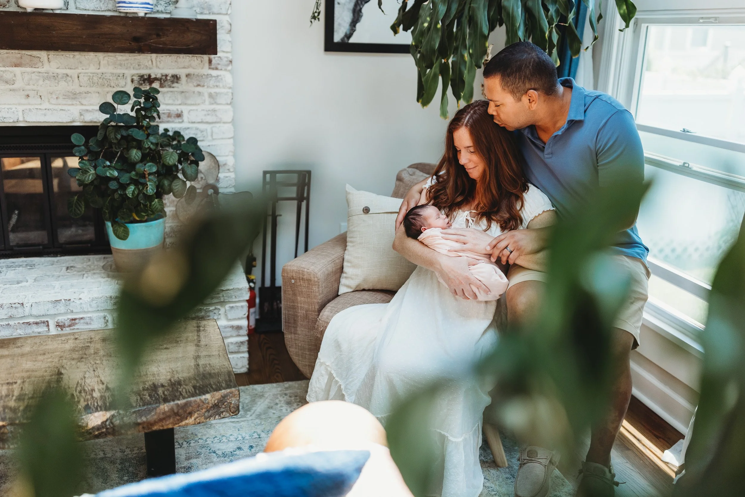 man kissing wife who is seated in a leather chair holding newborn baby and they are both looking at the baby