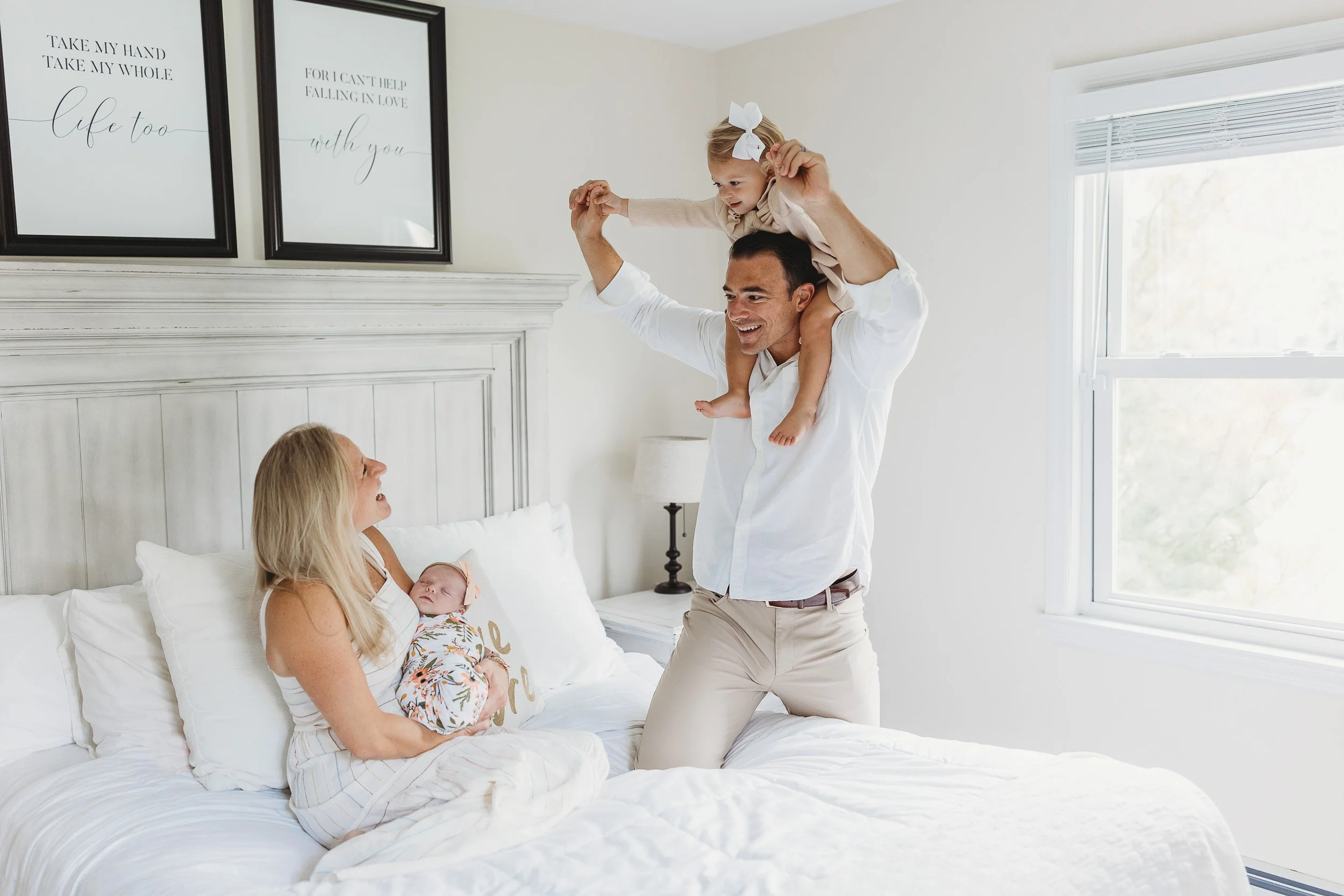 dad in his jacksonville home with daughter on shoulders walking towards bed and putting knee on bed while mom holds baby on her lap 