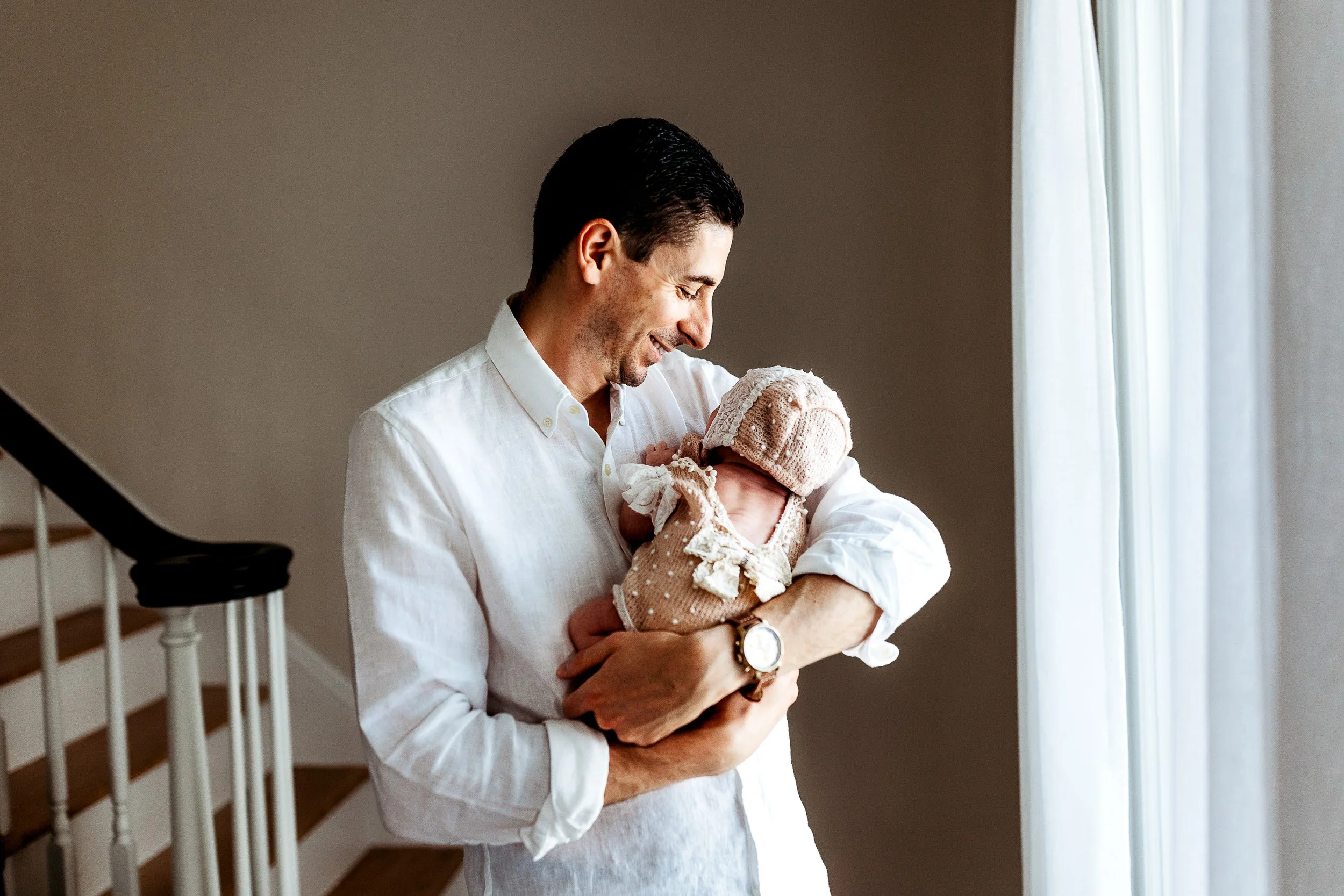 dad holding baby by an open door in a white linen shirt and jeans and baby in a vintage bonnet and knit onesie
