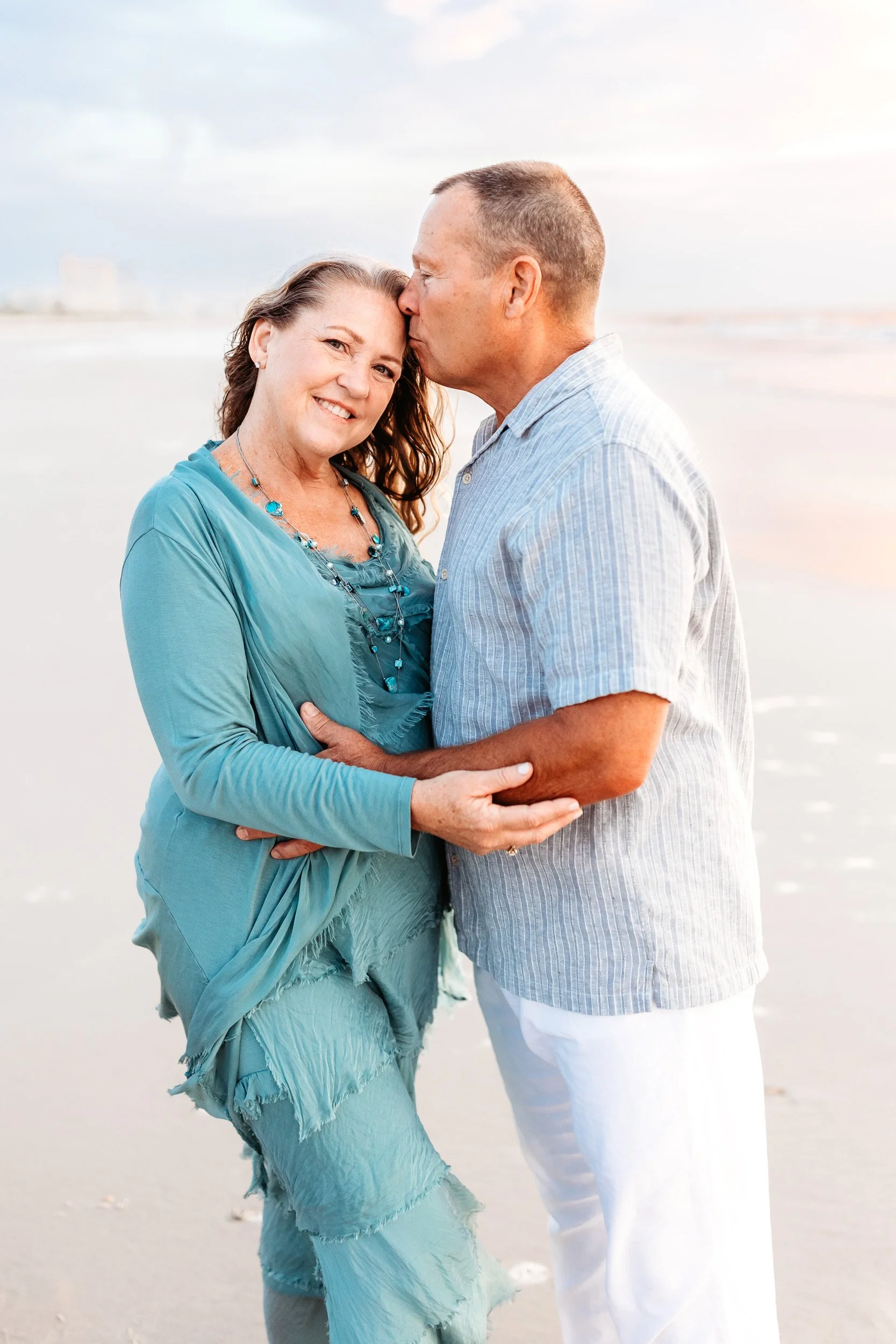 husband kissing wife on the head at sunrise on the beach in Jacksonville near the per