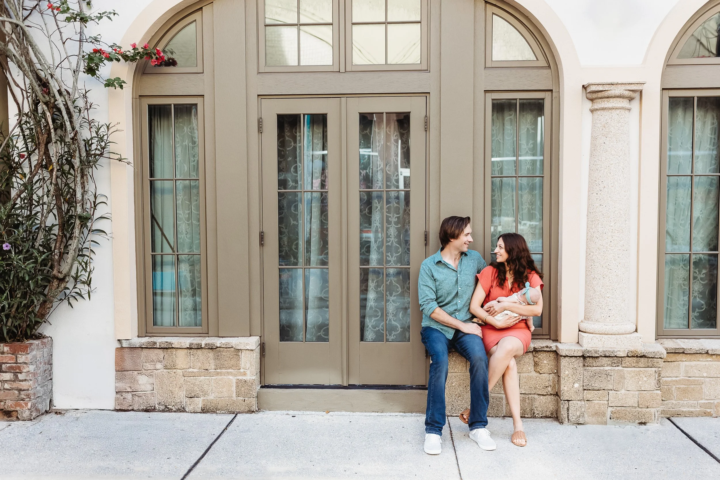 family seated on a ledge in front of a glass door with arch in historic area of st augustine and they are holding a baby 