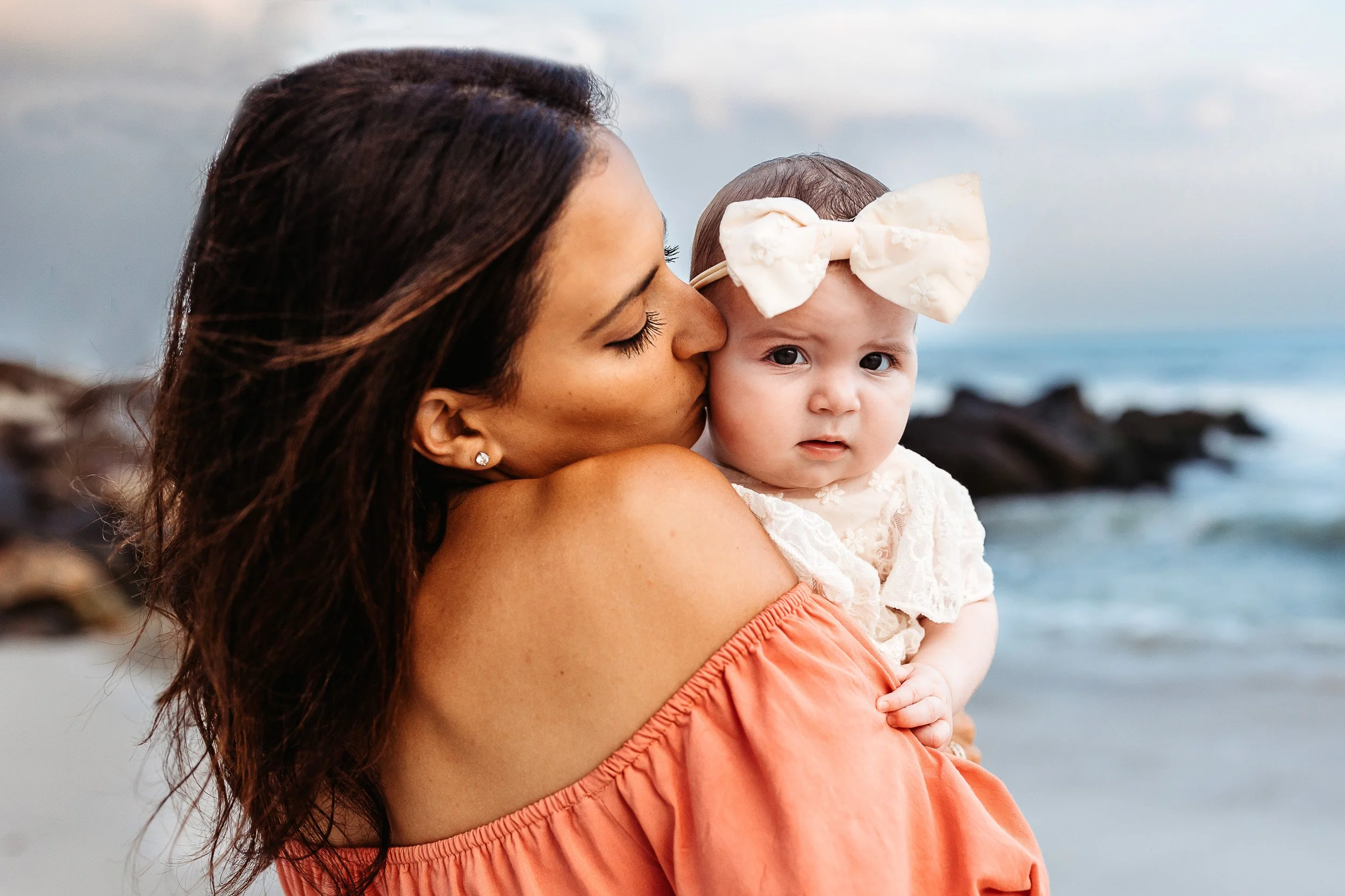 mother in coral off the shoulder dress kissing baby girl with large ivory bow on the beach in north eastern florida 