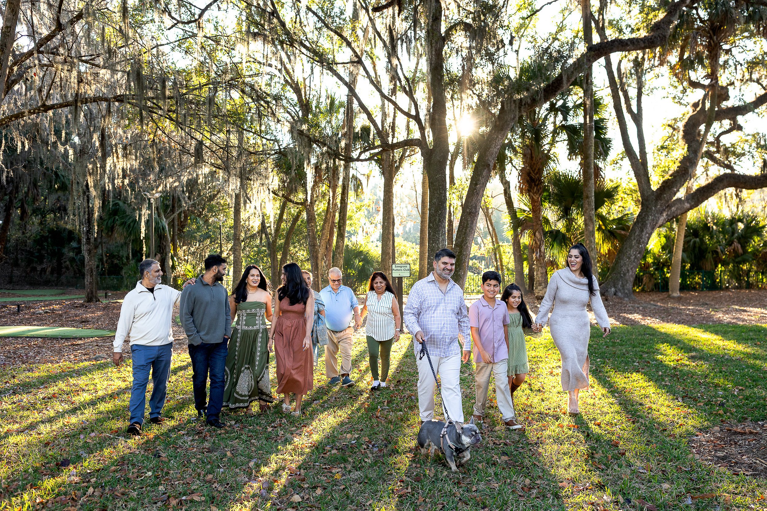family walking though sun drenched oak trees at sunrise through the sawgrass mariott and one man is walking a dog.  they are all laughing and holding hands 