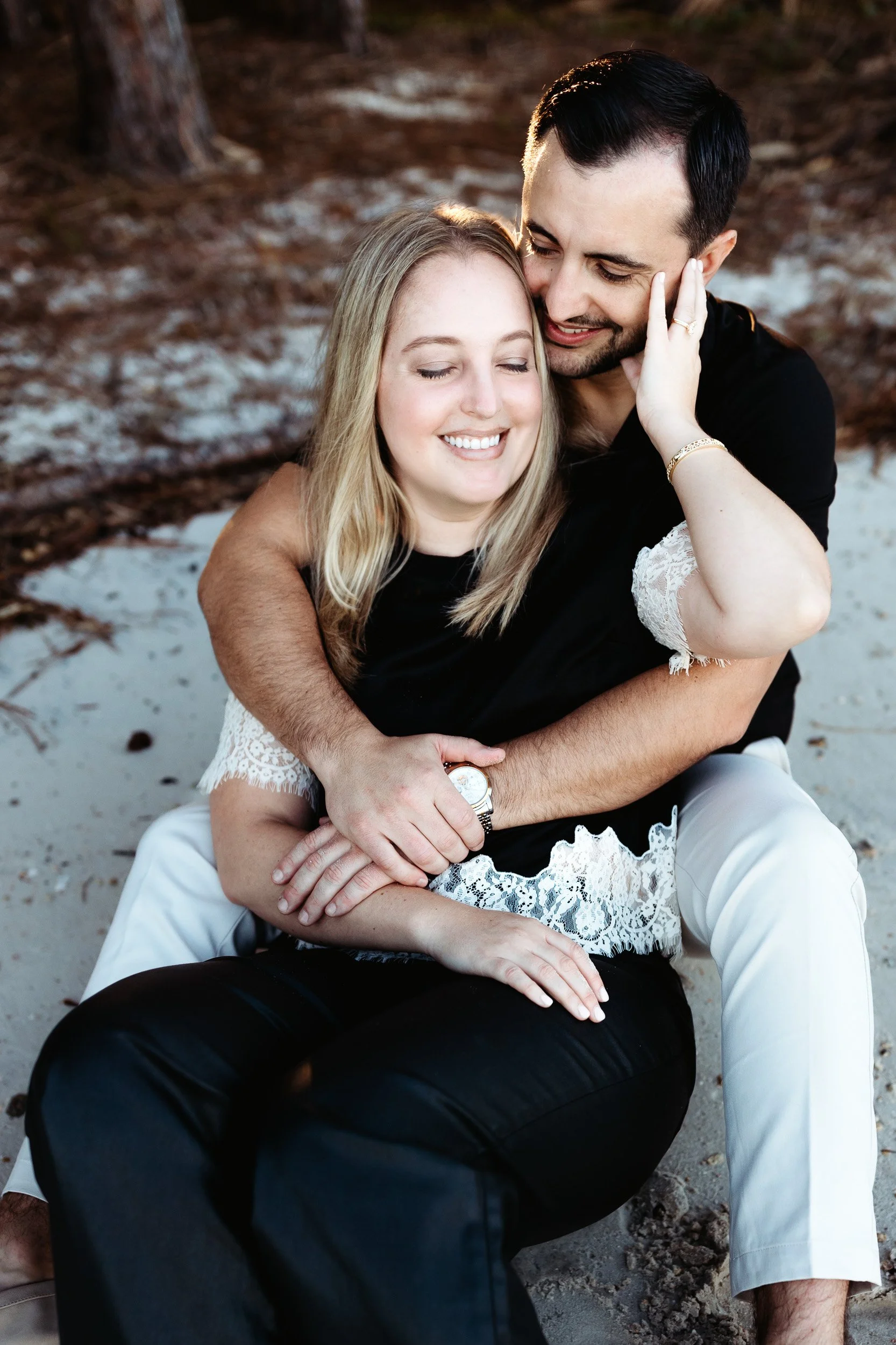 COUPLE embraced seated on the sand during nocatee engagement session and woman is touching man's face