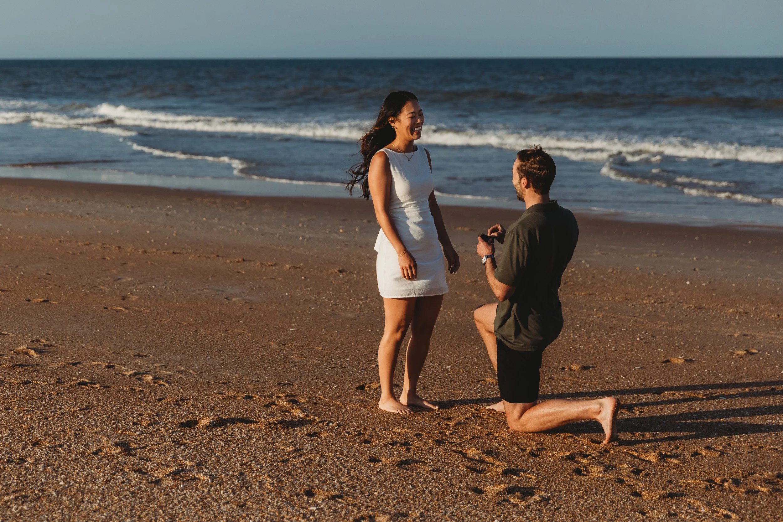 man on his knee proposing to girlfriend in front of the ocean at sunset