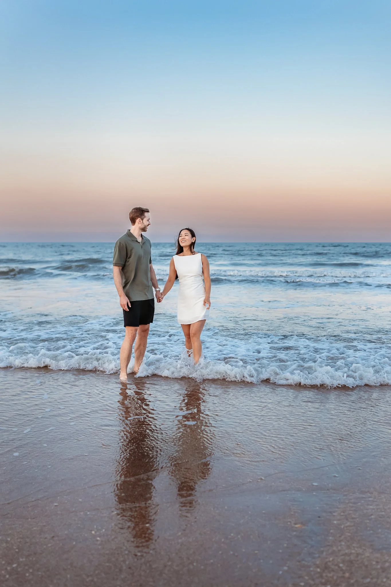 couple walking out of the ocean hand in hand with their reflections in water and pink and blue sunset sky