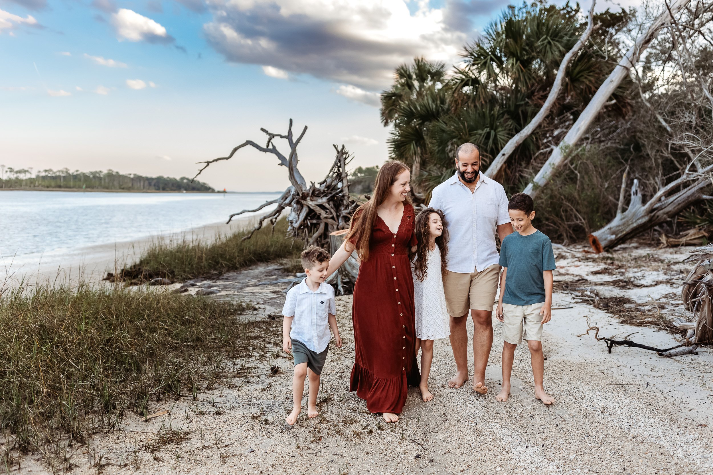 family walking along a beach in rust and teal outfits on the intracoastal for photos with Jacksonville photographer at sunset. they are laughing and looking at each other 