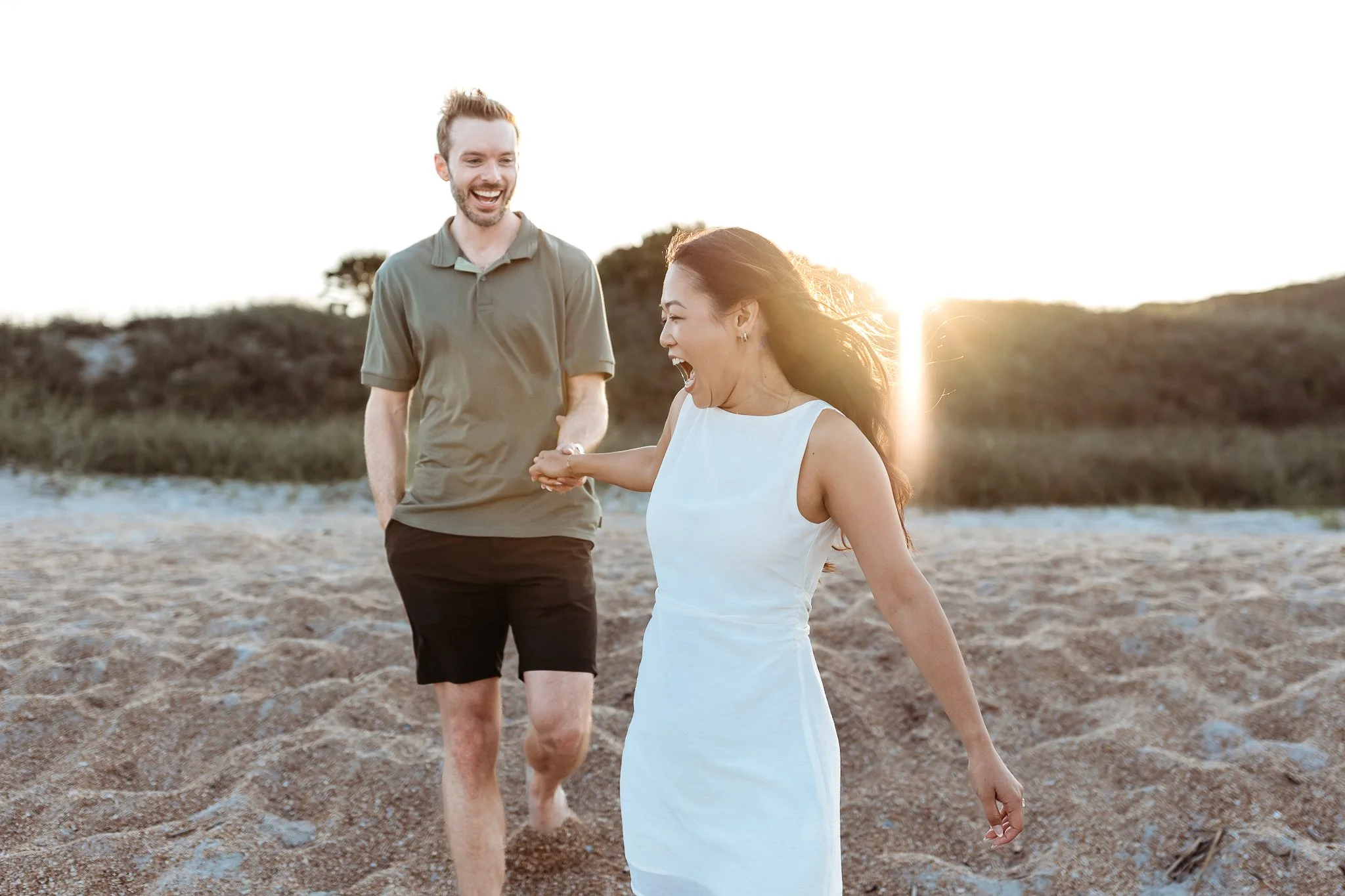 couple holding hands running down a hill at sunset on the beach in front of a dune and they are both laughing