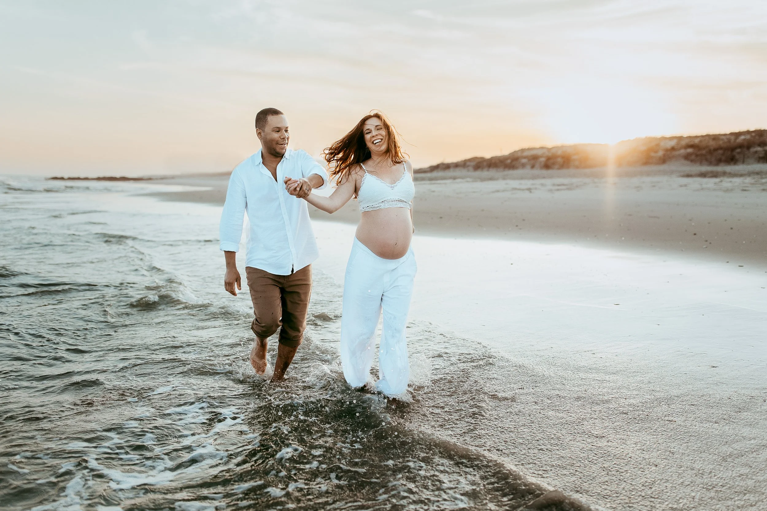 couple running down the ocean shore hand in hand and woman is pregnant