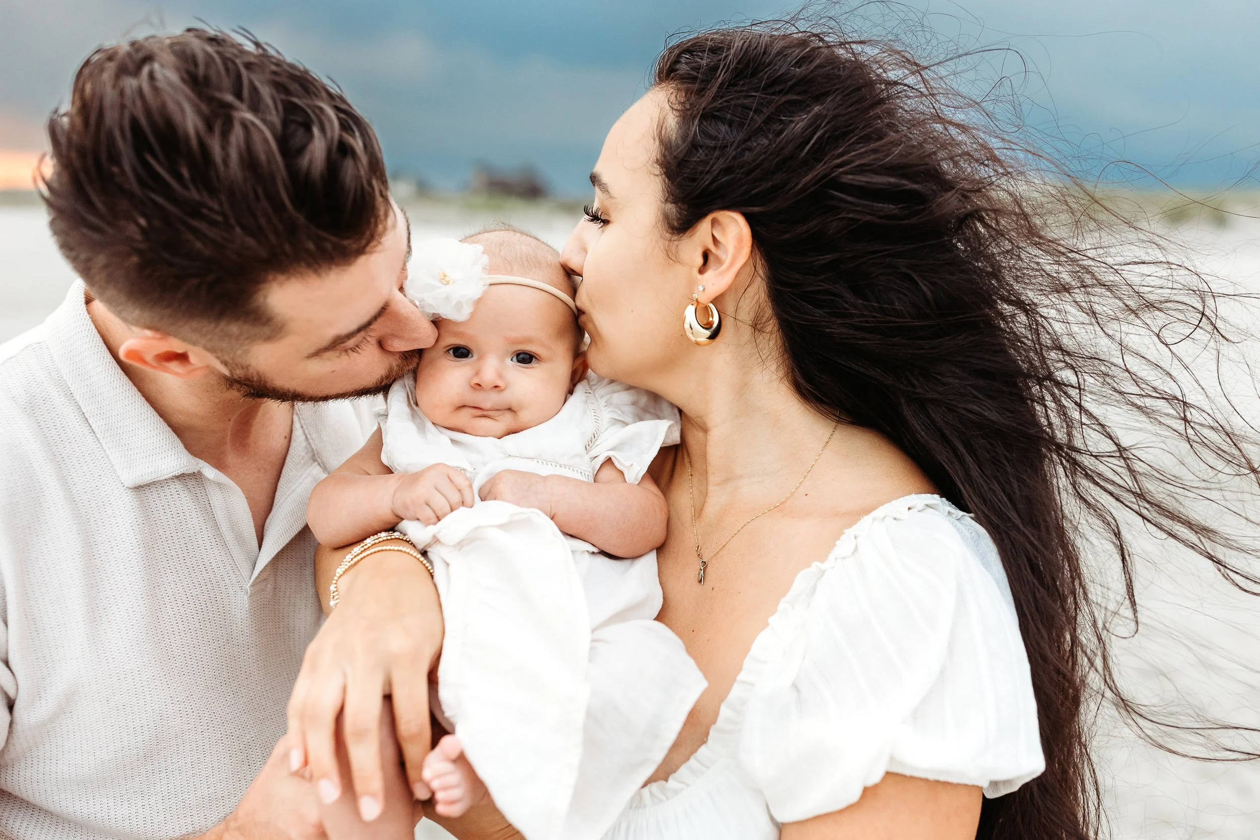 close up of parents kissing a baby who is looking straight at the camera and they are on the beach at sunset