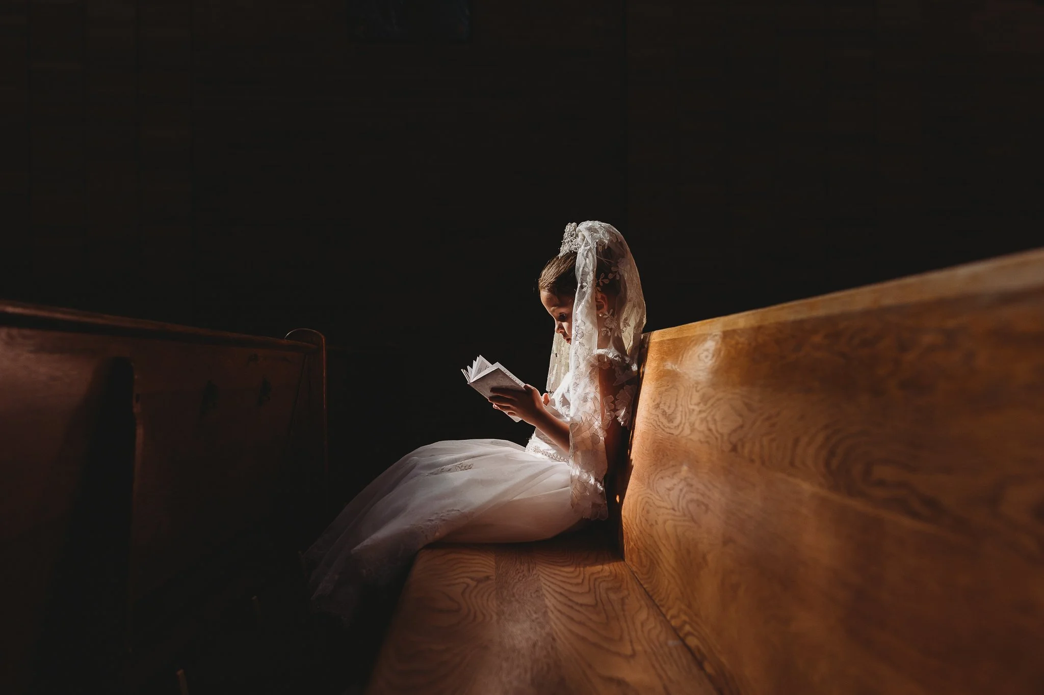 girl in communion dress in a pew at church reading bible with sliver of light on her
