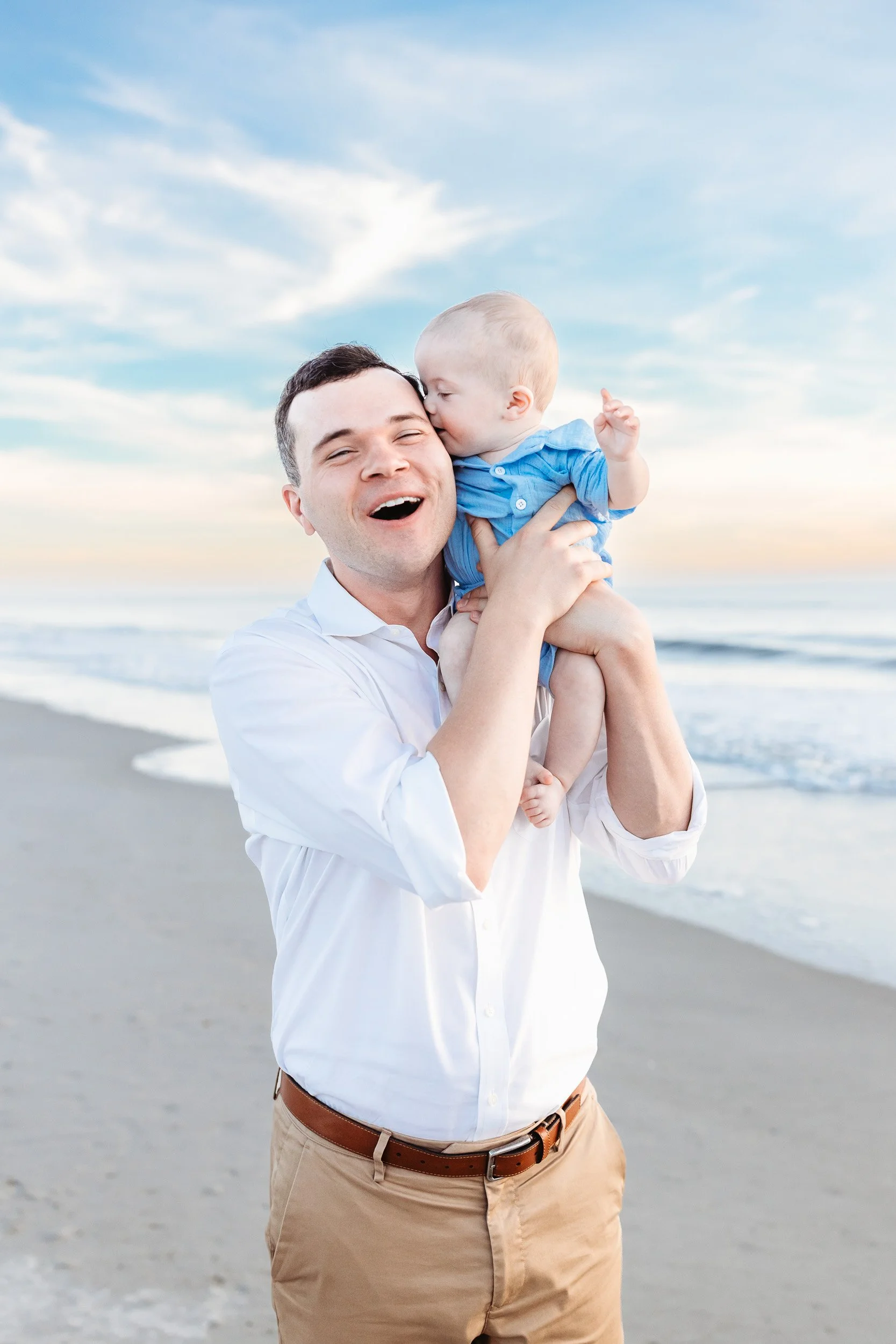 baby boy on dad's shoulder and kissing dad on cheek and dad is laughing