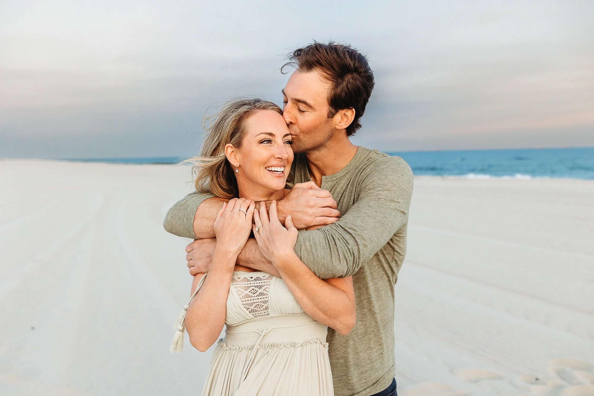 couple on the beach in Jacksonville and husband is kissing the wife on the head and she is laughing they are taking photos with their family