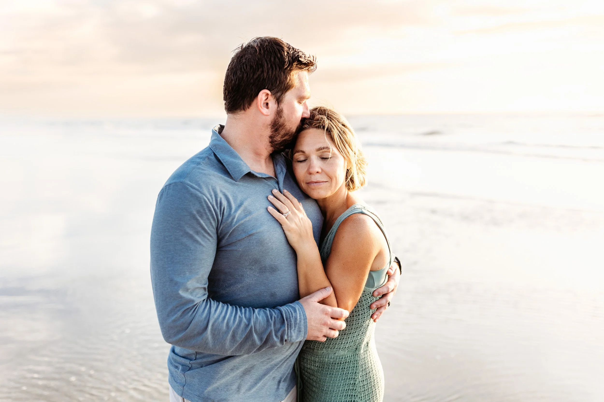 husband kissing his wife and she is leaning against his chest at sunrise on the beach