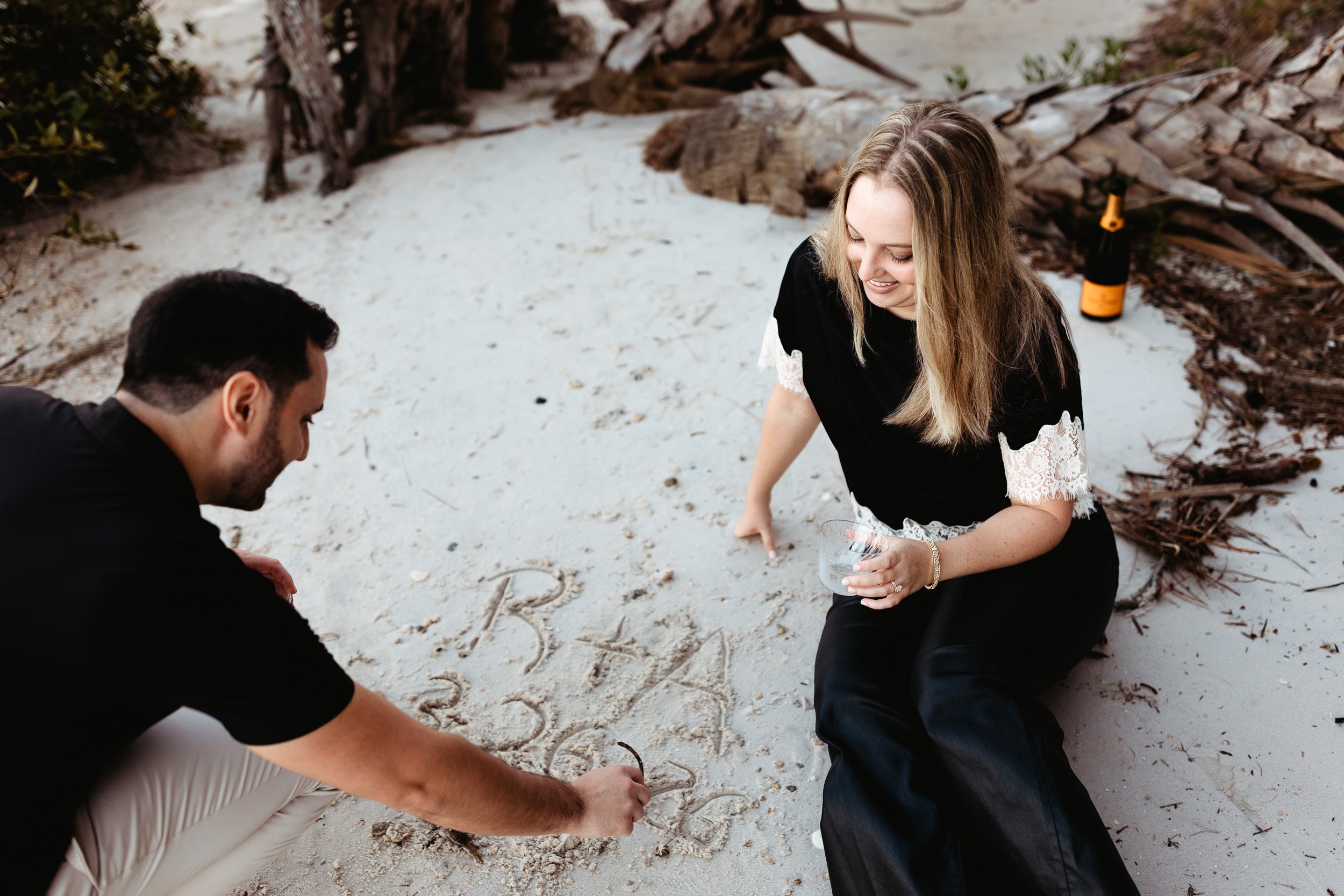 mAN drawing in the sand while girlfriend watches during an engagement photo session