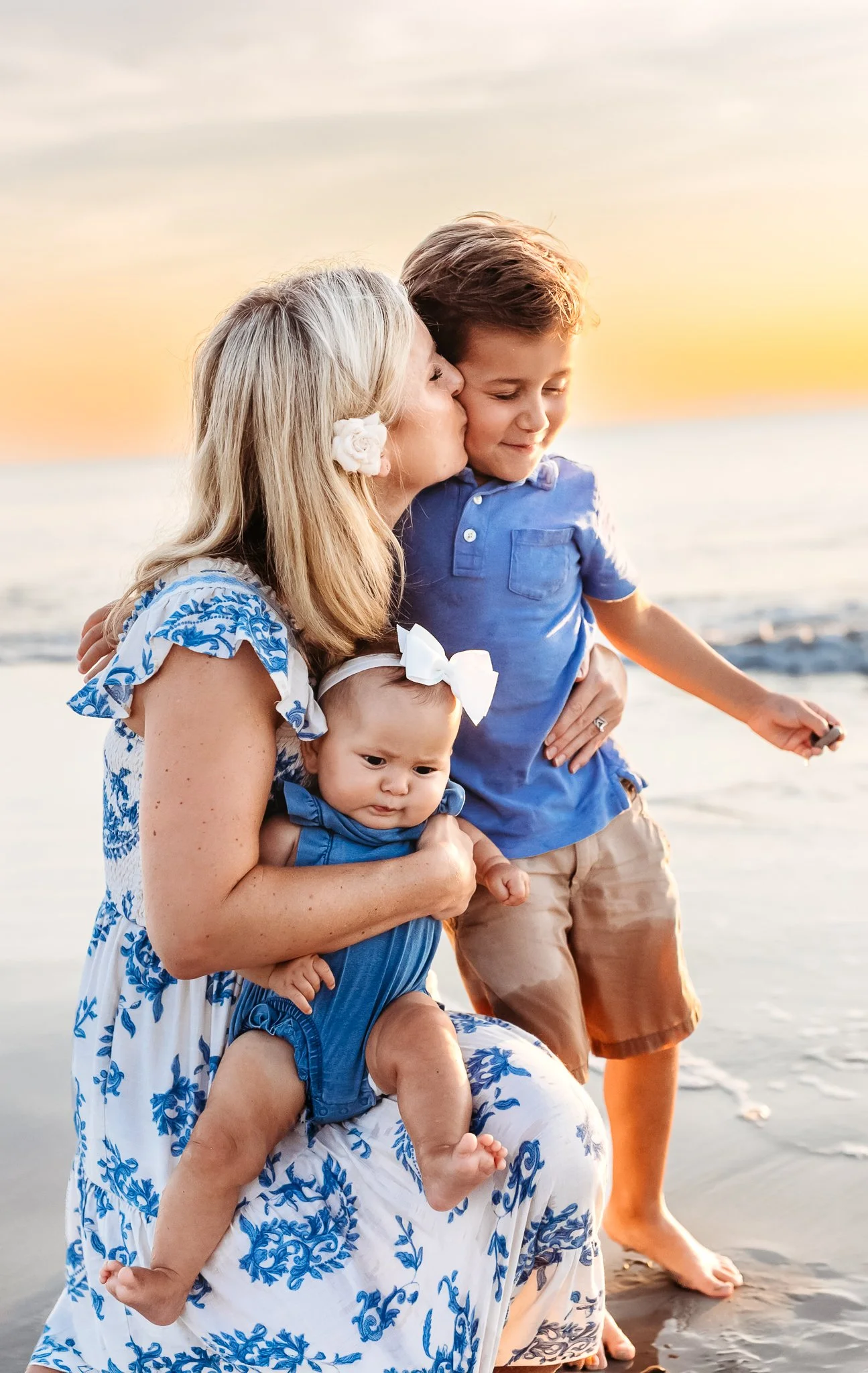 mother kneeling on the water's edge at the beach at sunrise holding a baby and kissing her son