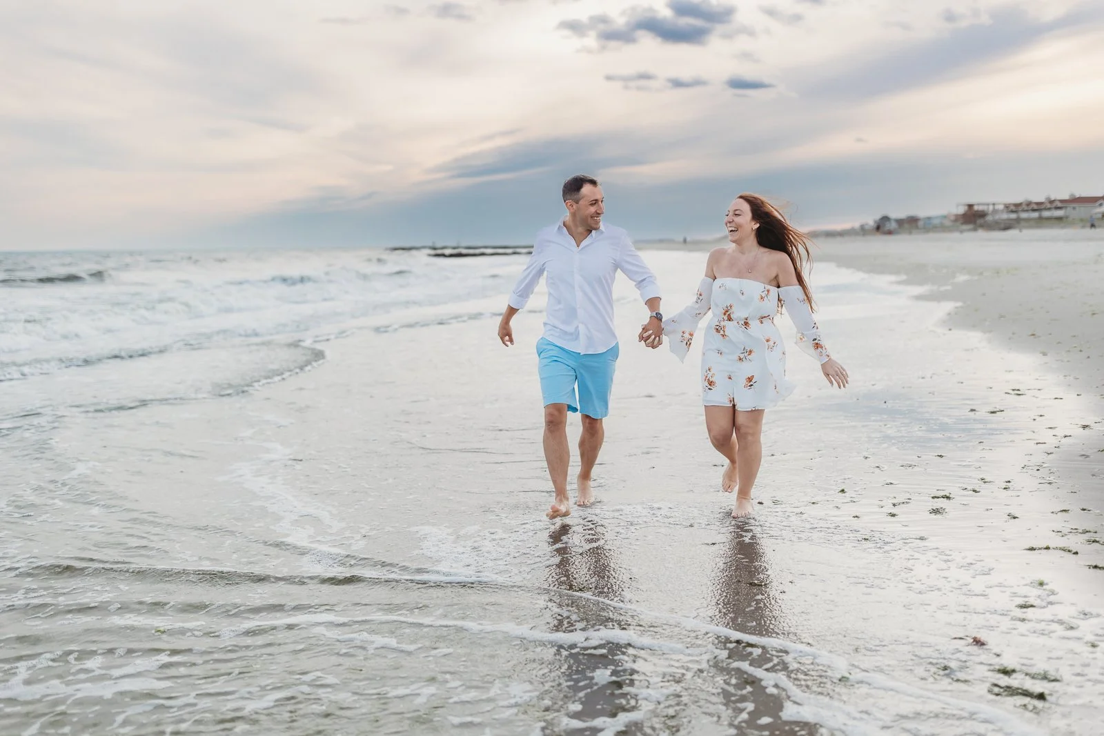 man and woman holding hands running along shore of ocean aftr getting engaged and they are laughing and it is sunset