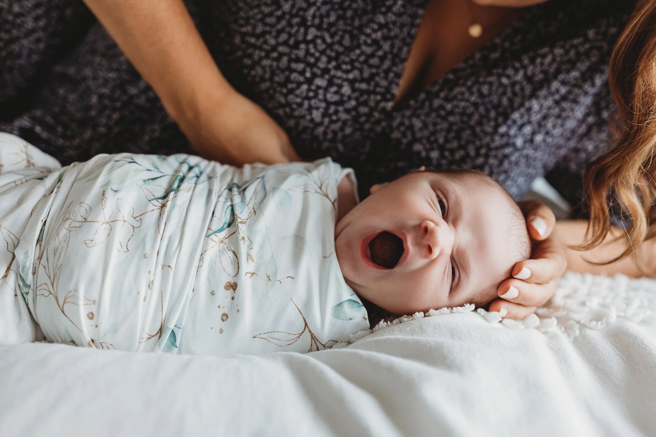 baby swaddled in a jersey knit blanket mom's hands on head lying on bed baby is yawning
