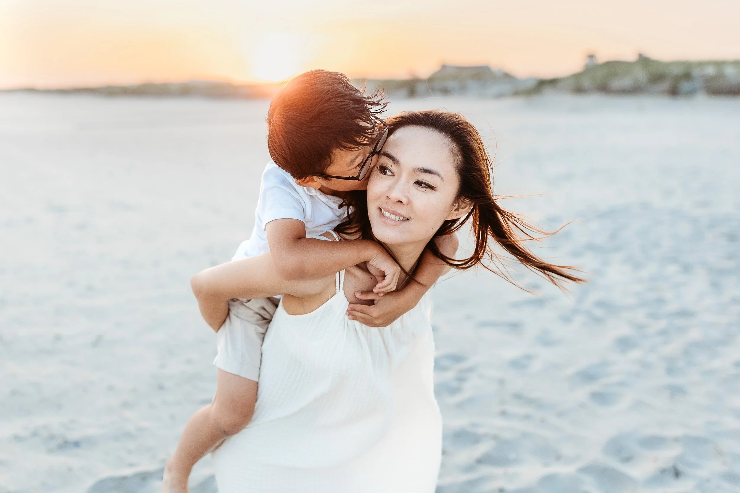 little boy on mom's back and he is kissing her while the sun sets behind them on the beach